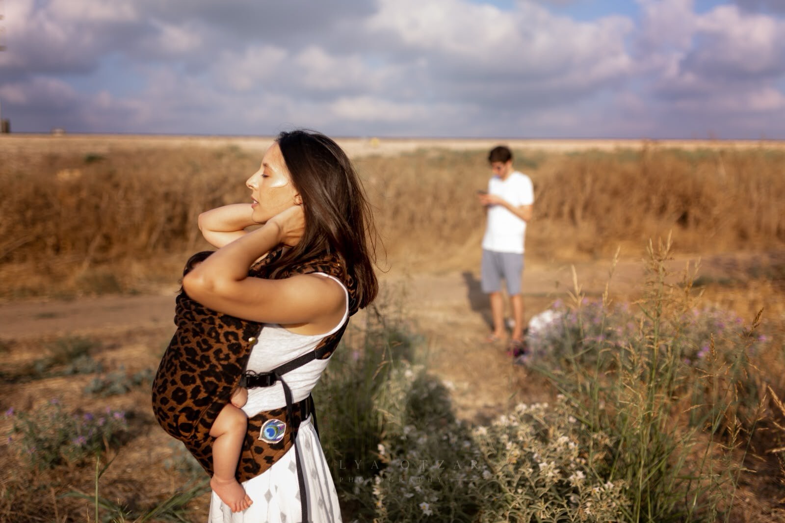 A mother carrying her baby in a sling across an open field, the father walking behind