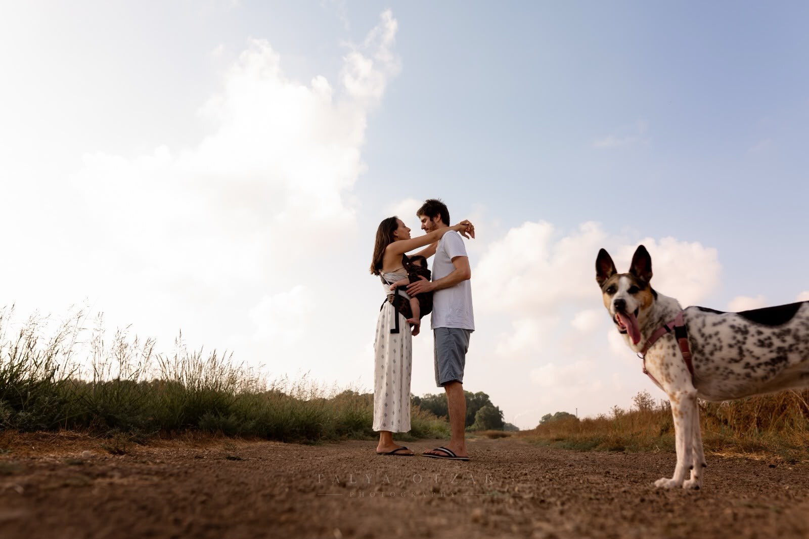 Parents lifting their baby between them on a track under a wide sky, the family dog in the foreground