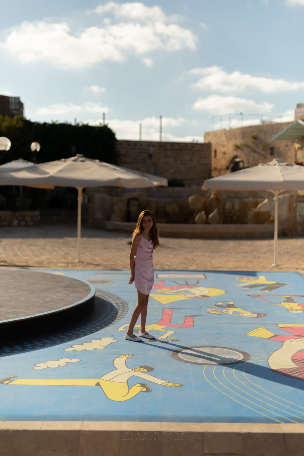 A girl standing alone on a huge painted mural on the ground, parasols and stone buildings behind her