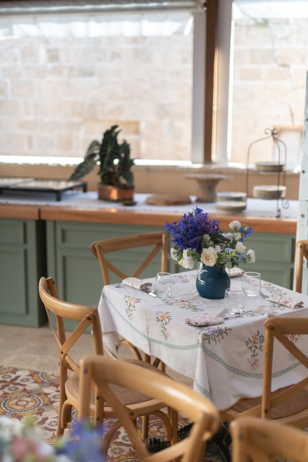 A table laid with an embroidered cloth and a blue jug of irises in a green country kitchen