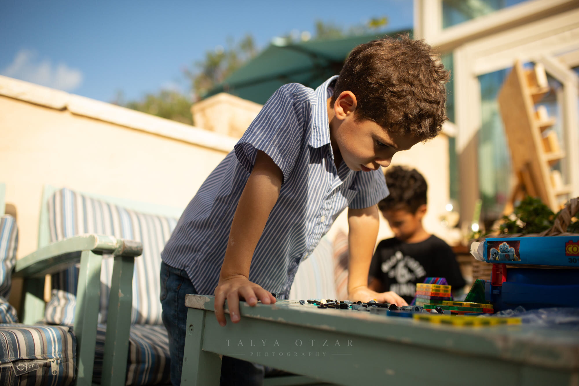 A boy leaning over a garden table absorbed in his toy cars, his brother playing behind