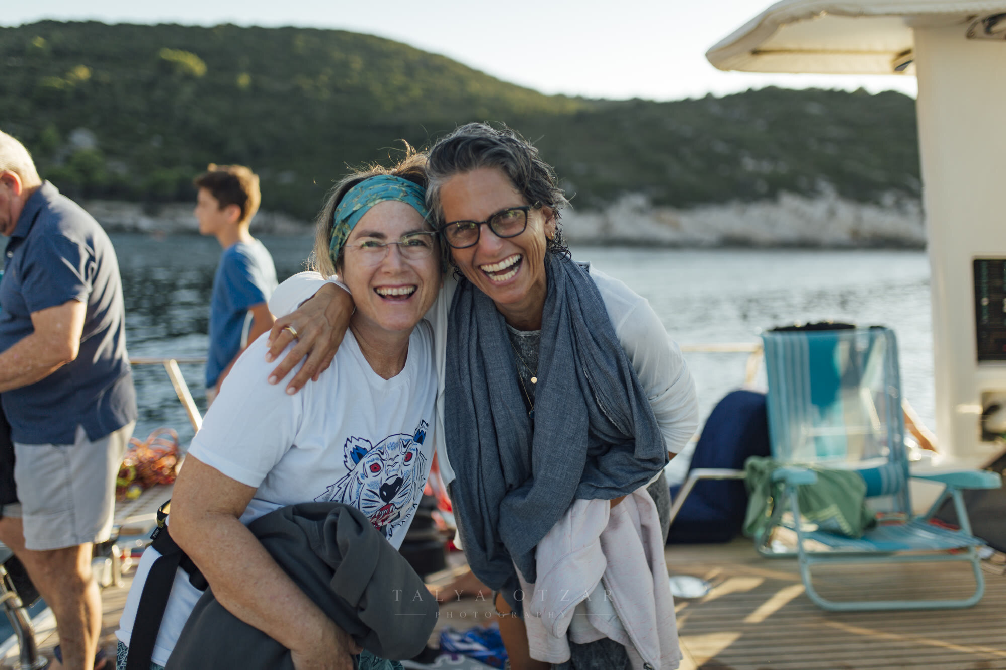 Two women laughing arm in arm on a boat deck, a green headland behind them