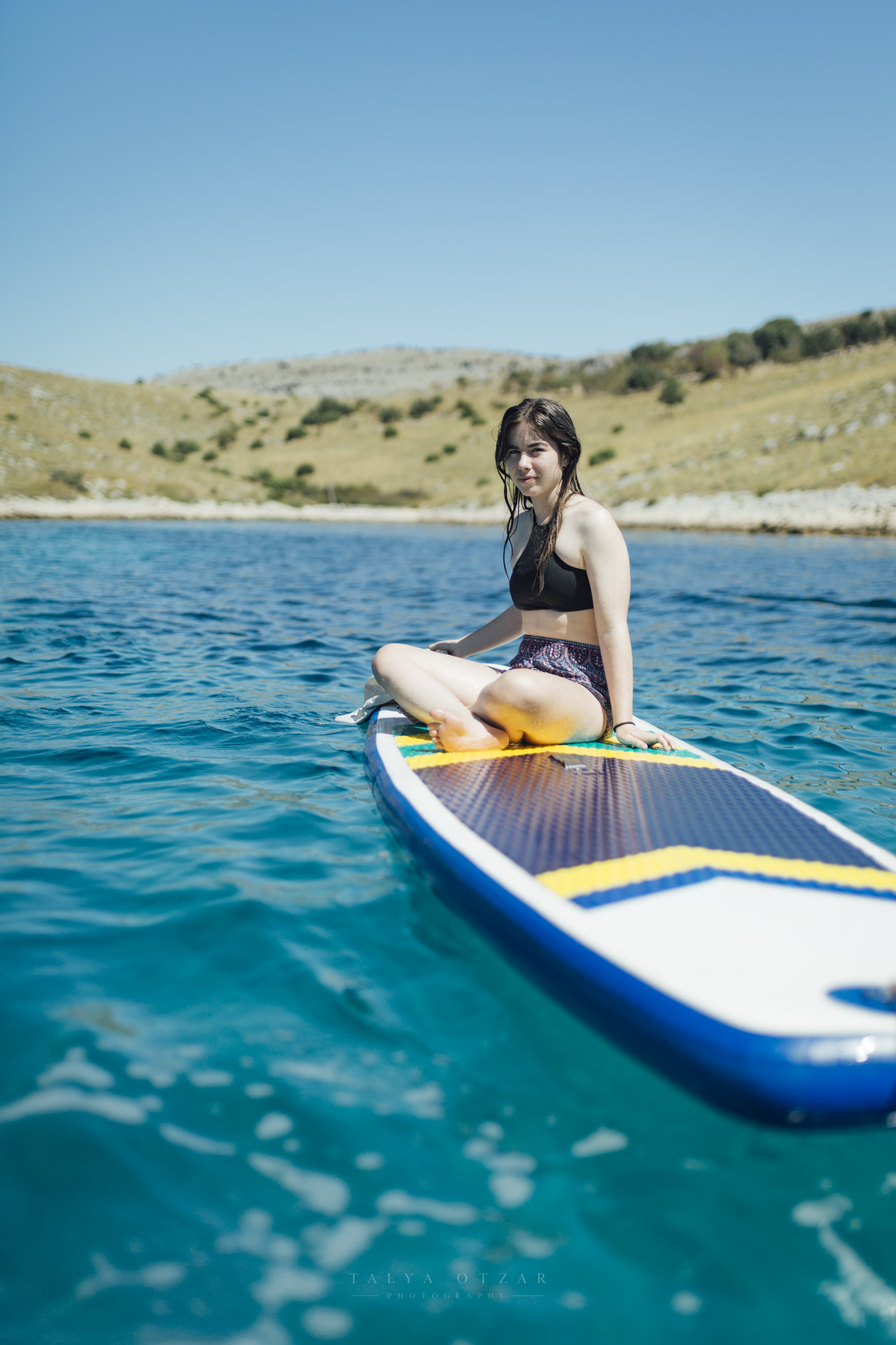 A girl sitting on a paddleboard on clear turquoise water, a bare hillside behind