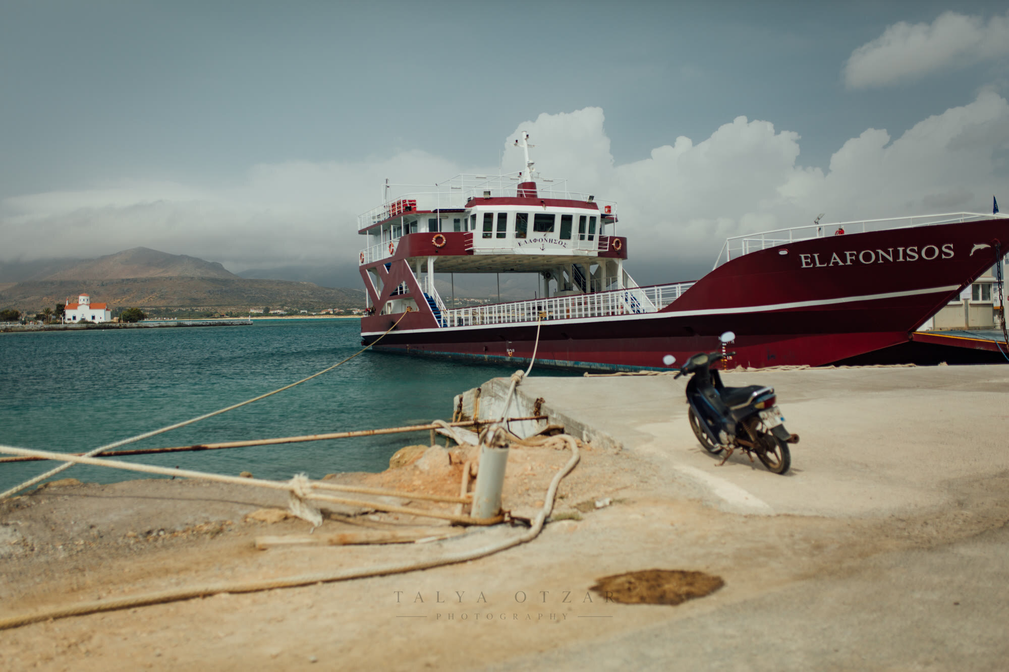 The island ferry tied up at the quay, a moped parked alongside and a white chapel across the water