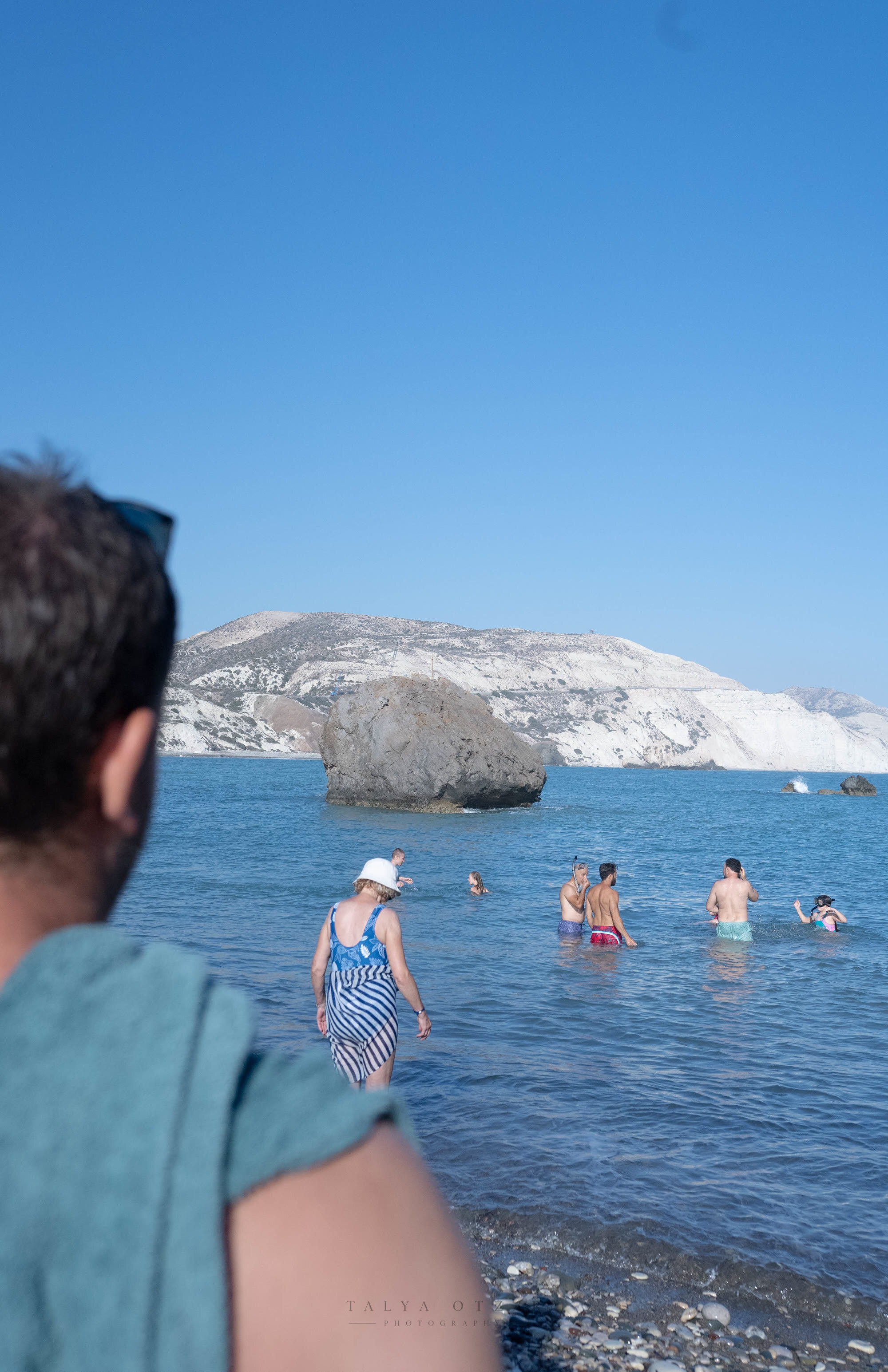 Swimmers in the shallows seen past a shoulder in the foreground, white cliffs behind