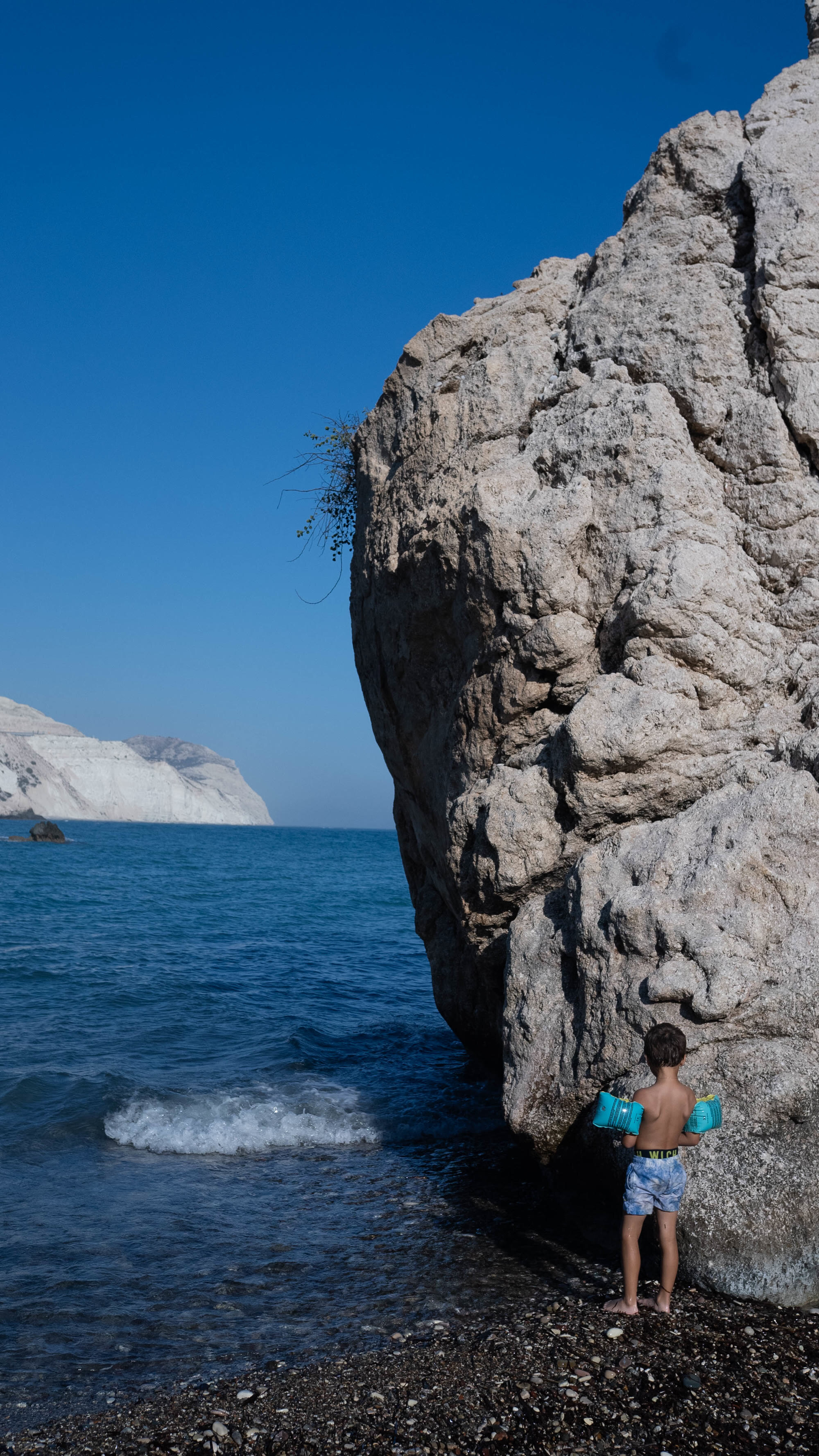 A small boy in armbands standing alone at the foot of an enormous sea rock