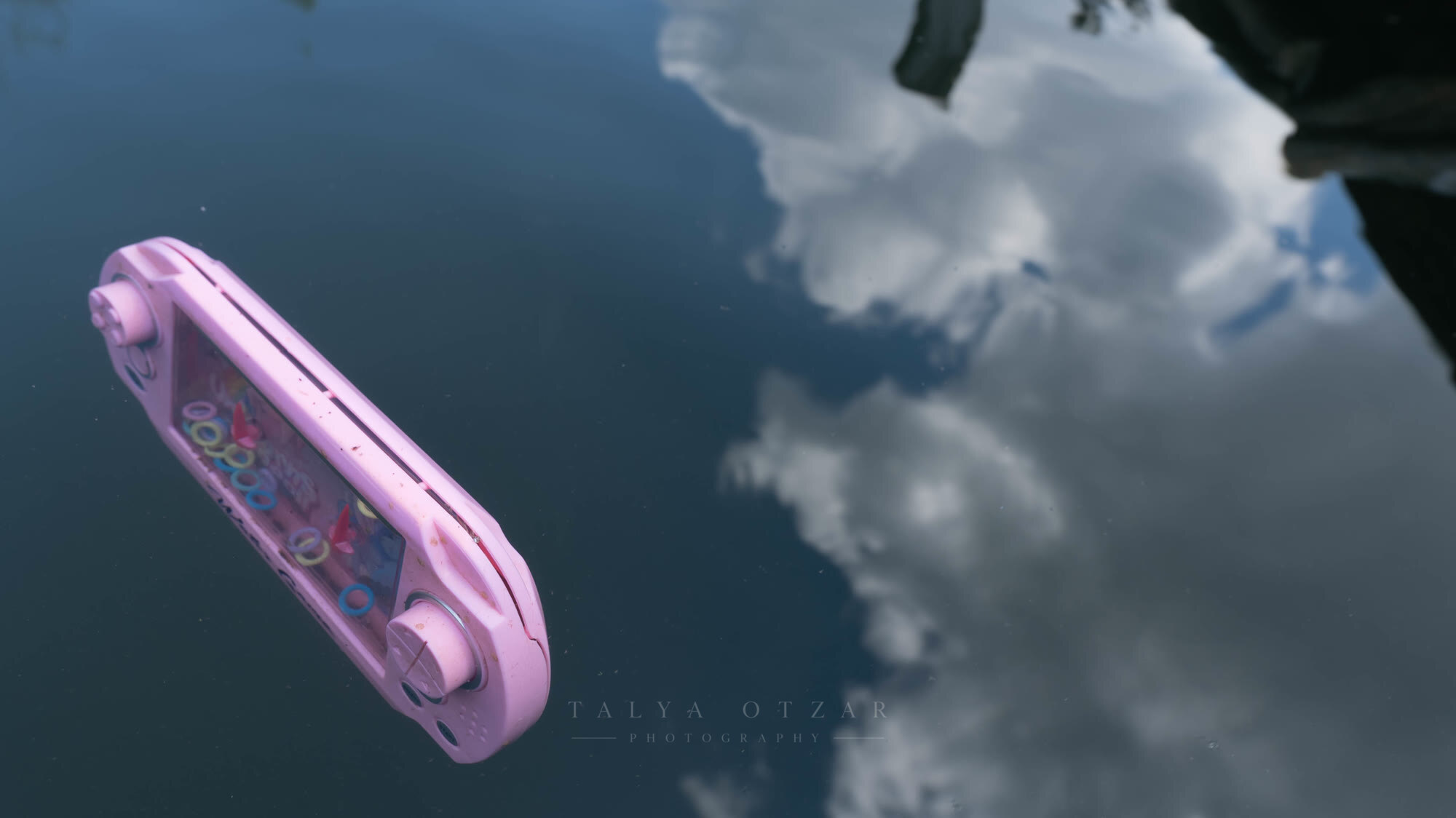 A pink handheld game console left on a wet car roof reflecting the clouds