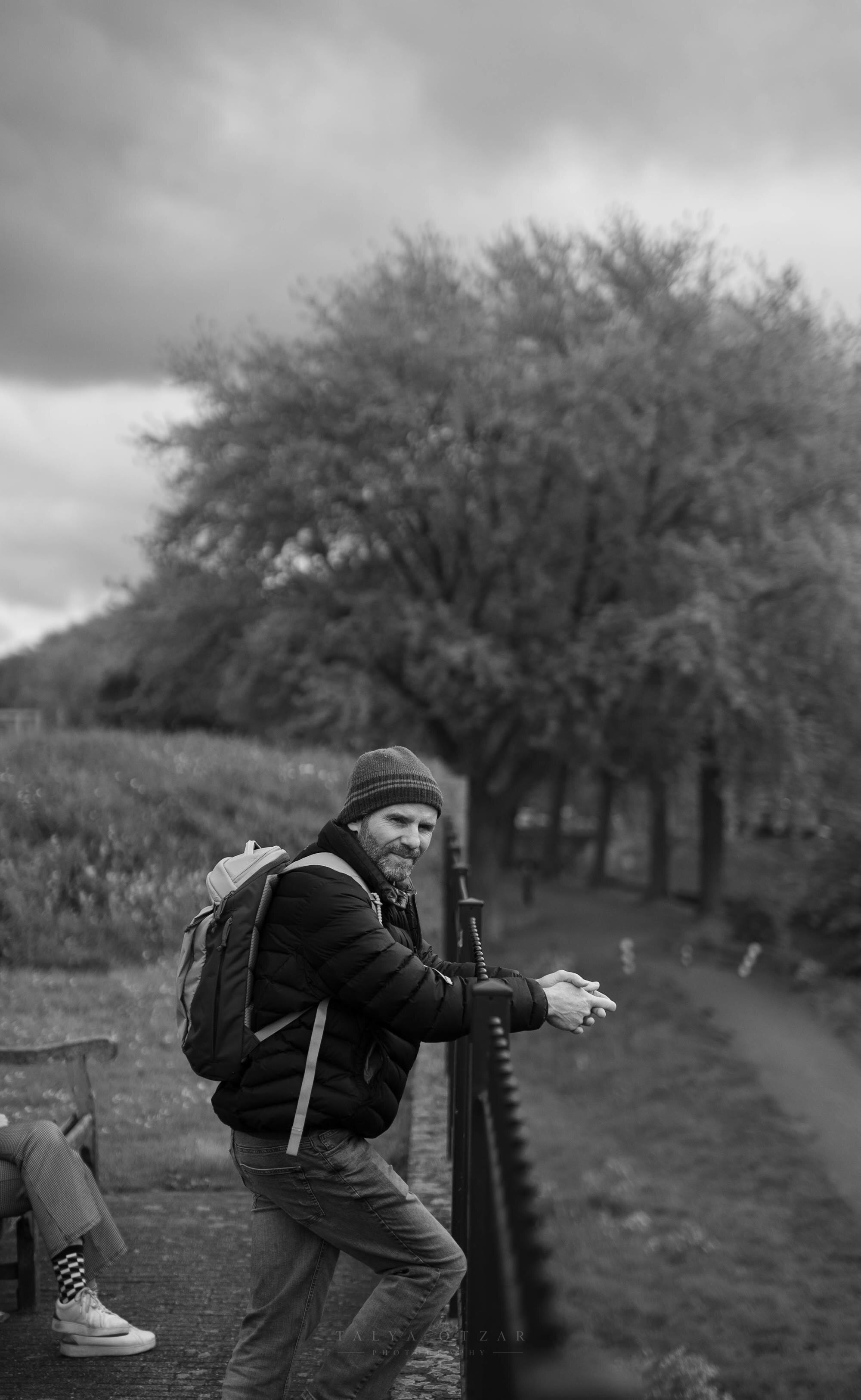 A man in a beanie leaning on the railing of a tree-lined path under a heavy sky