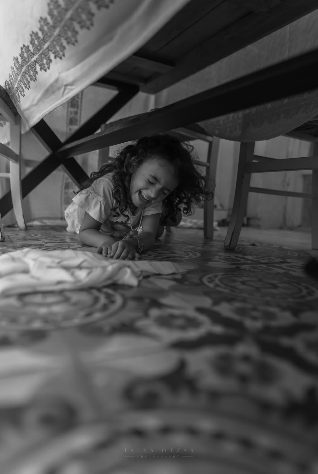 A small girl lying laughing on the patterned tiles under the table