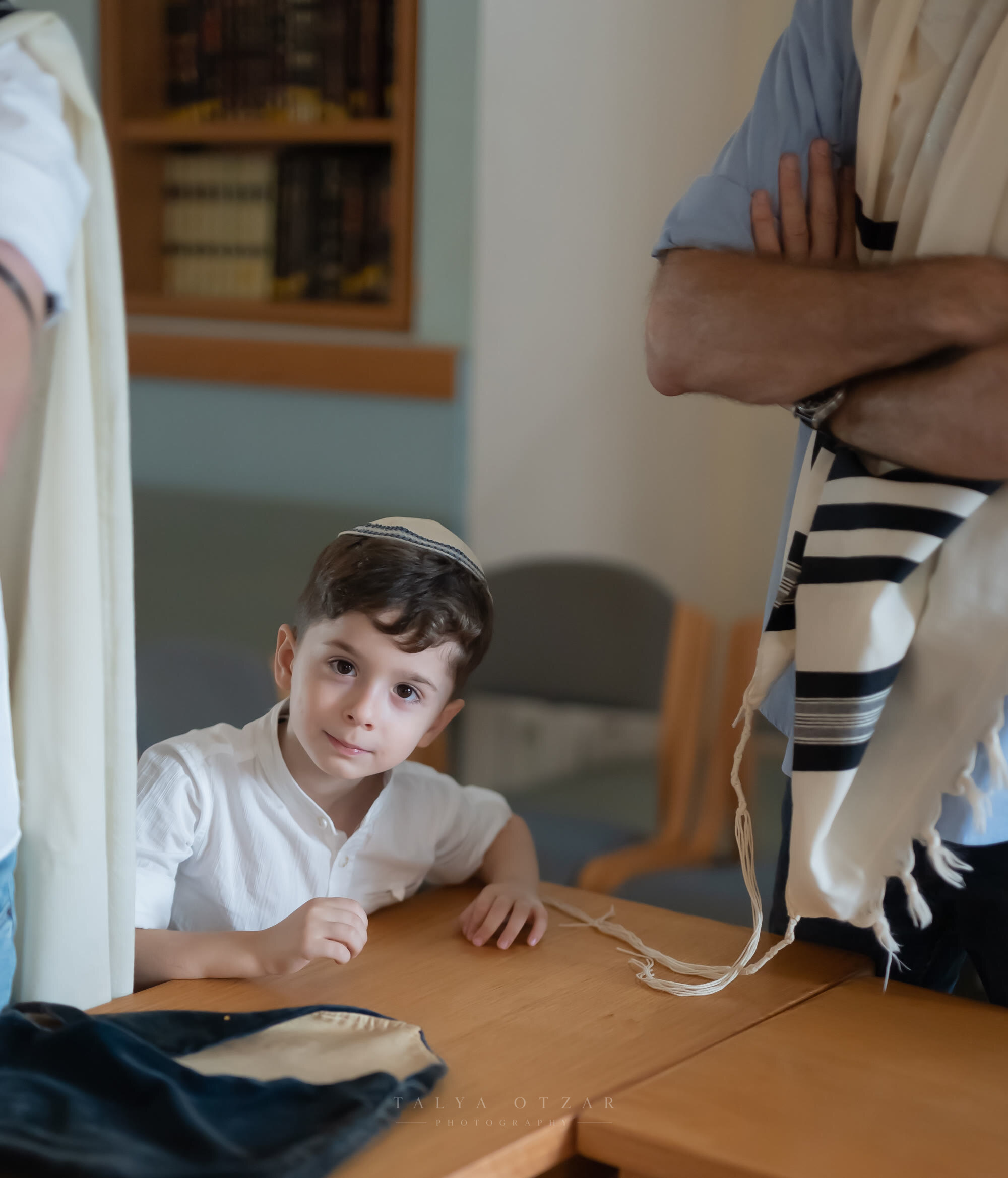 A small boy in a kippah at the table watching his father put on a tallit