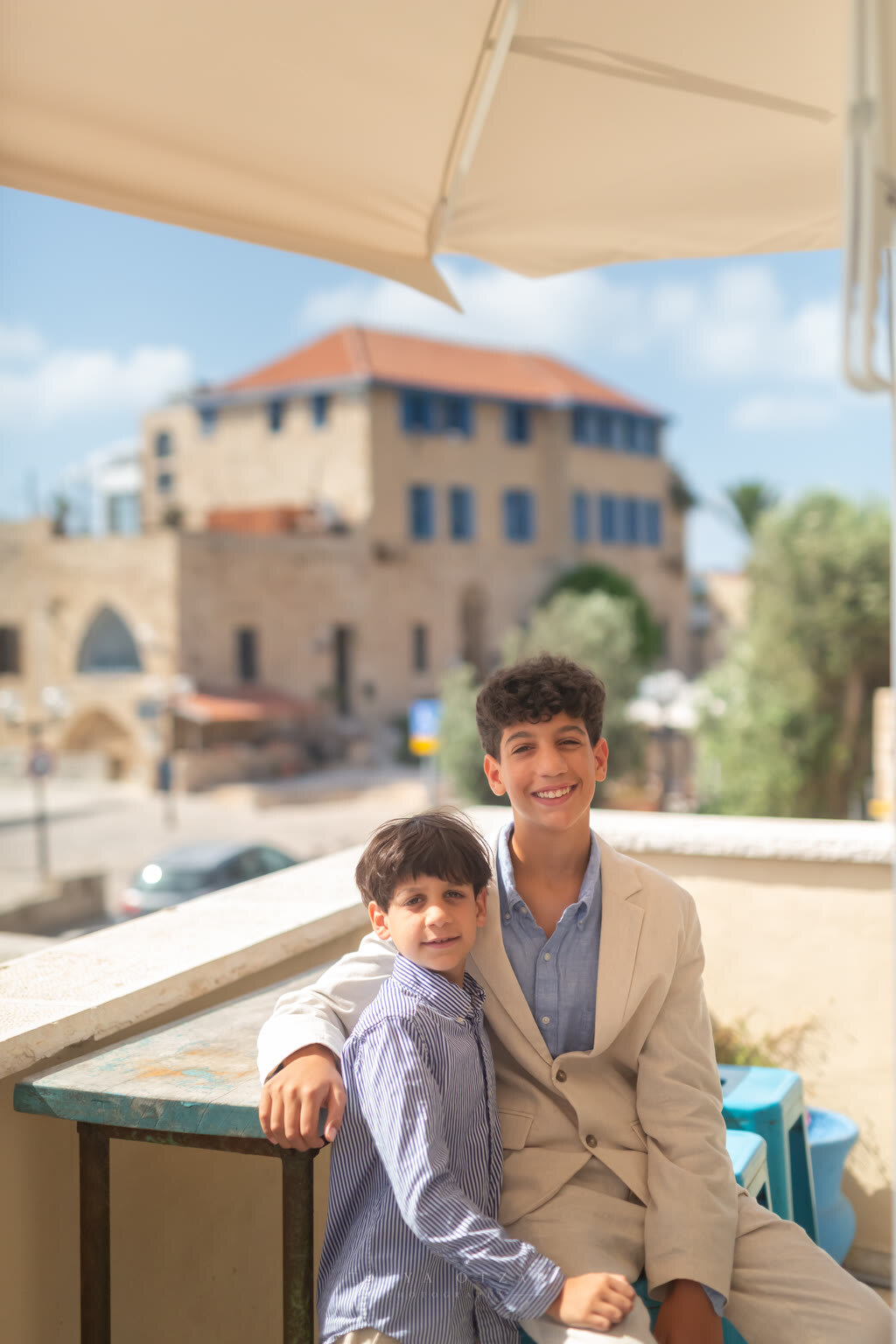 Two brothers on a Jaffa terrace, the older in a suit, an old stone house with blue shutters behind