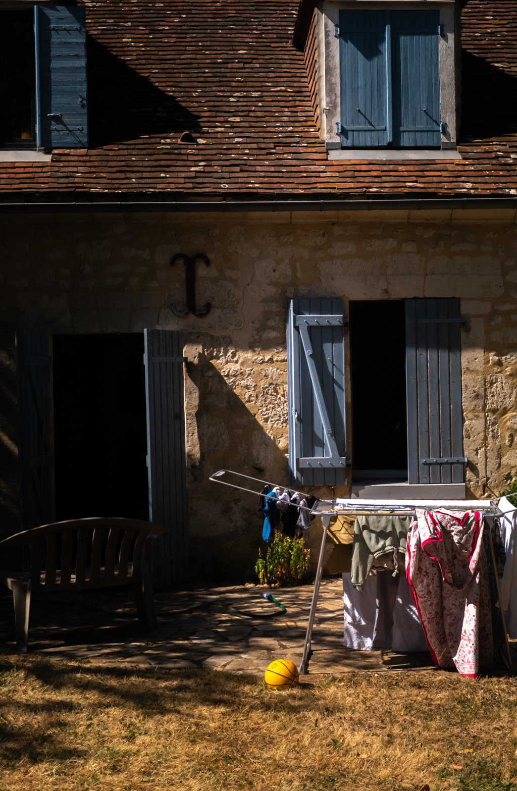 Washing on an airer outside a shuttered stone farmhouse, a yellow ball on the grass