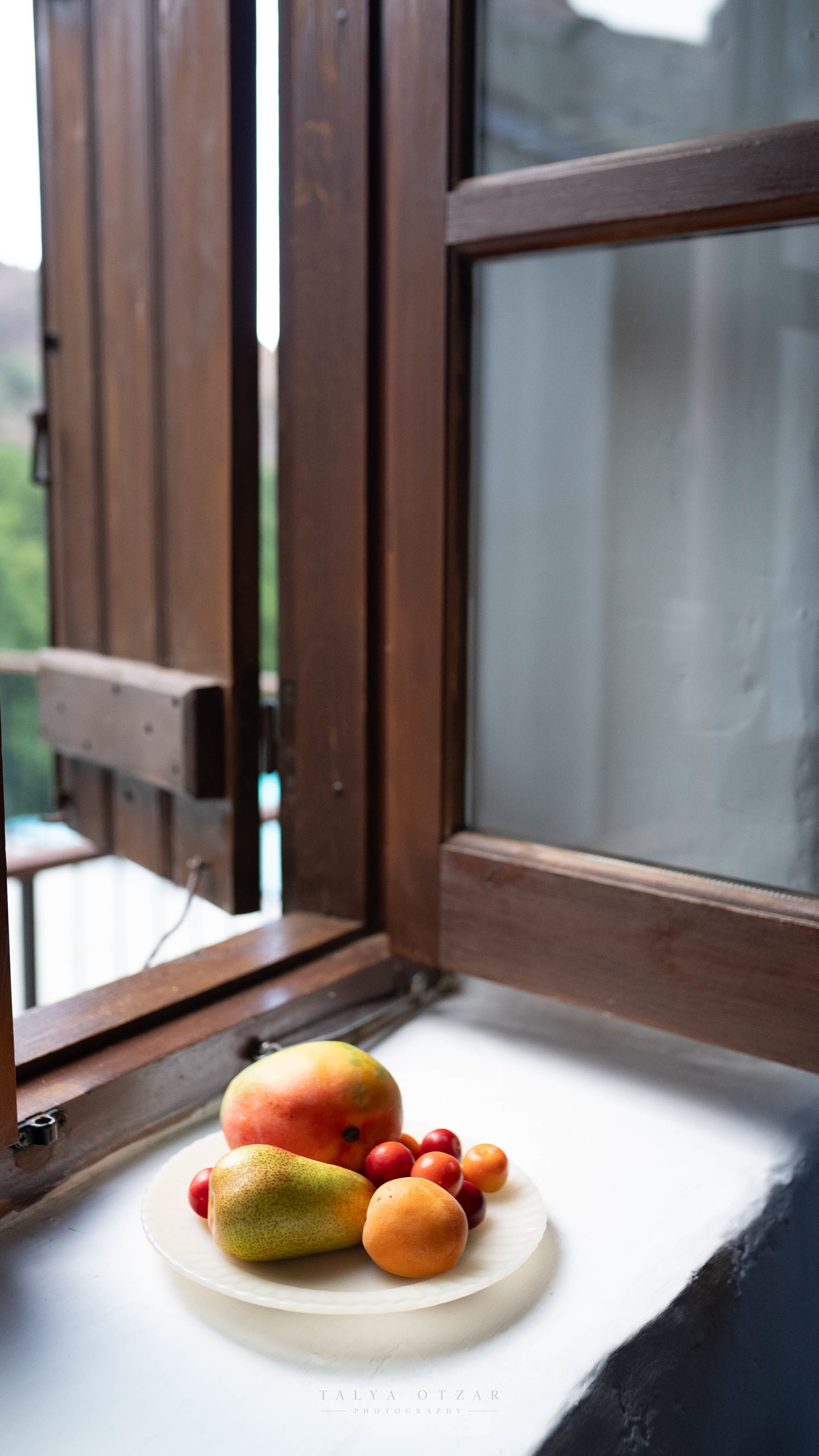 A plate of mango, pear, apricots and plums on the windowsill by an open shutter