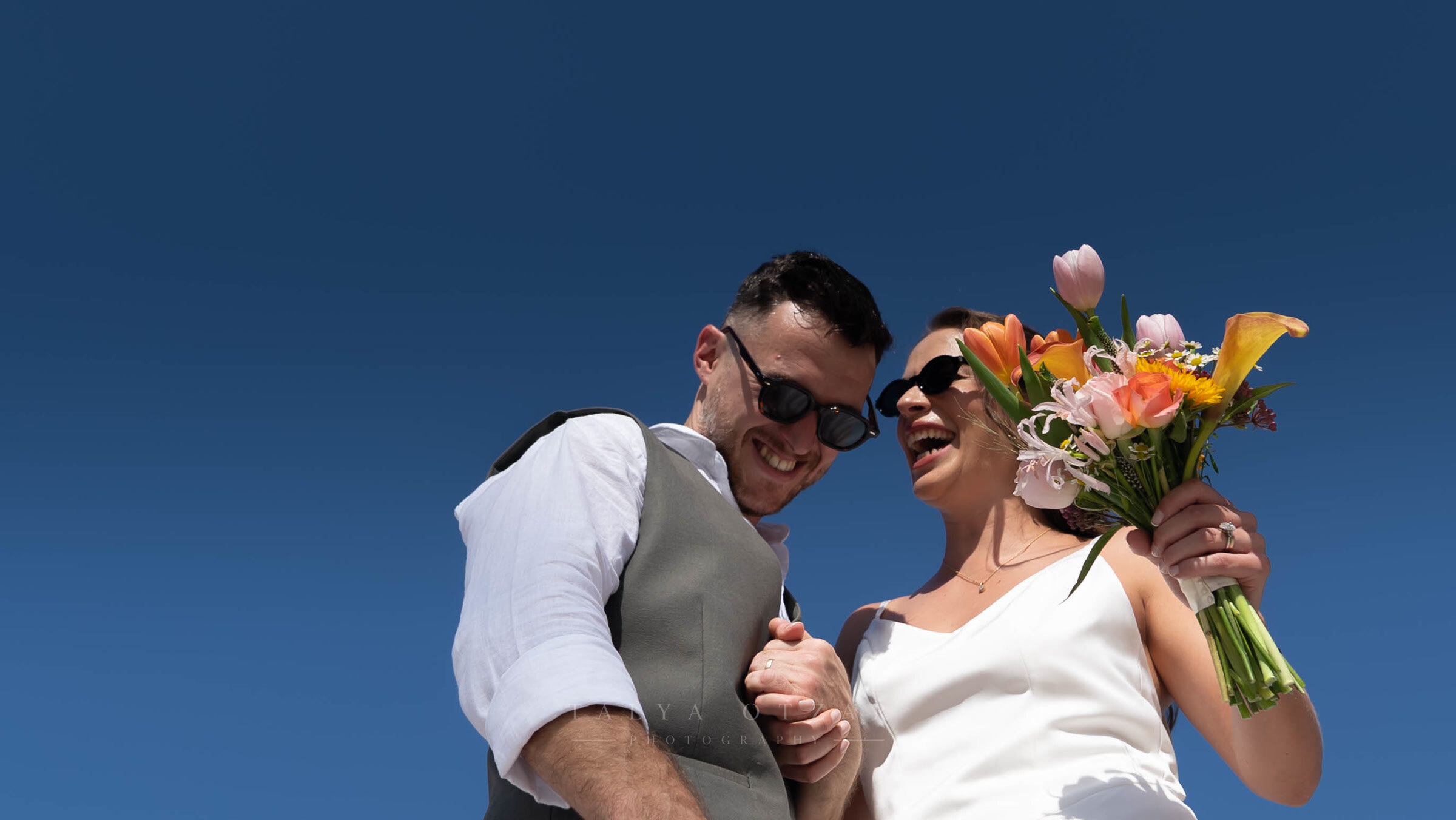 Bride and groom in sunglasses laughing together against a deep blue sky, the bride holding a bouquet of tulips and calla lilies