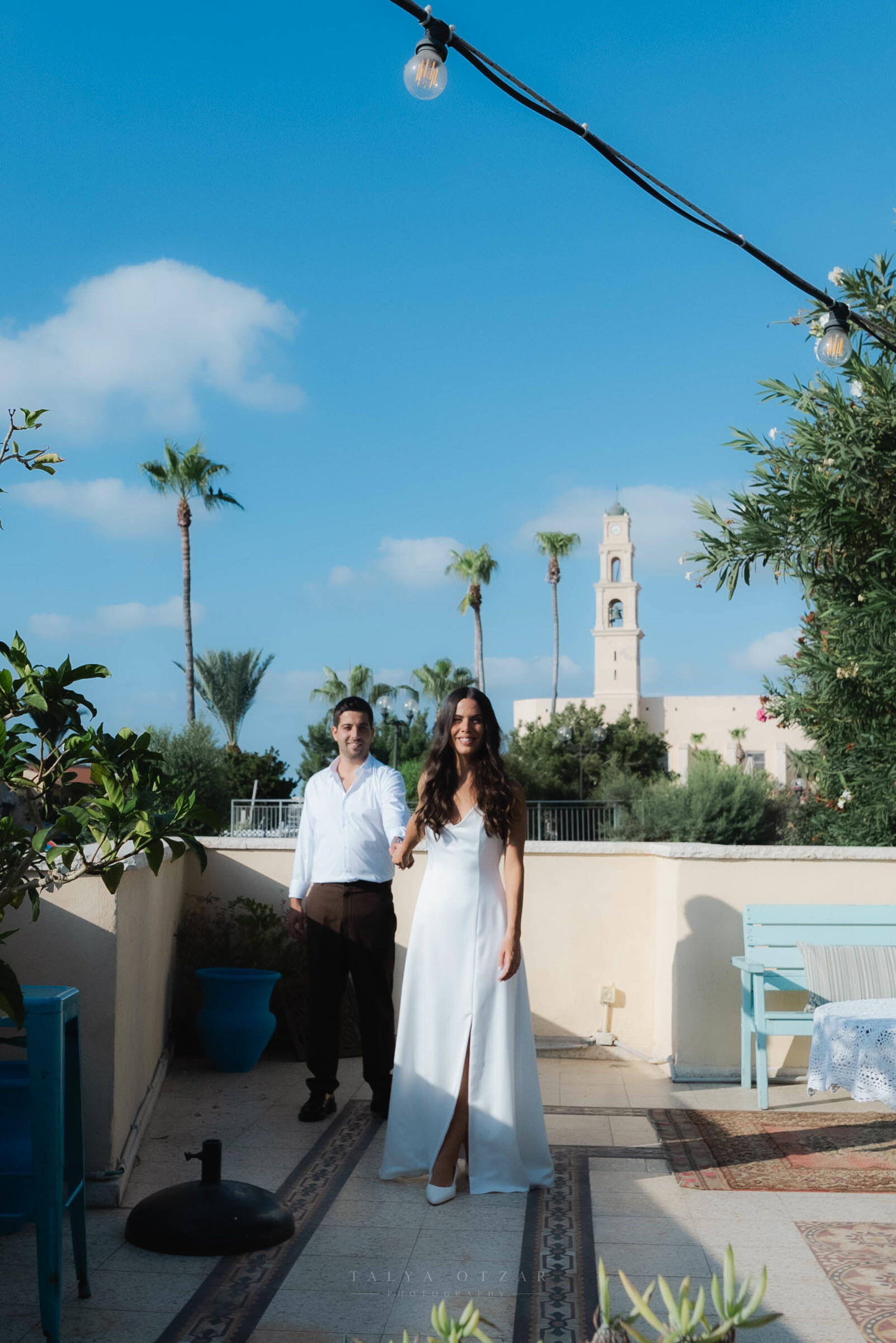 Bride in a satin gown and groom on a Jaffa rooftop terrace, the bell tower of St Peter's Church and palm trees behind them