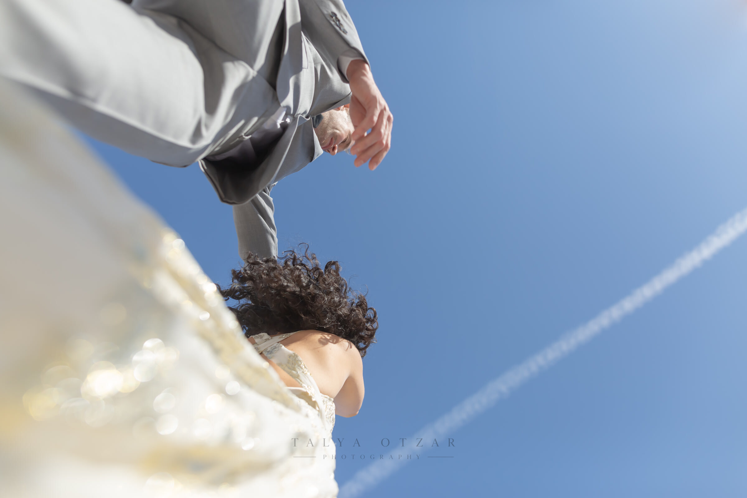 Low-angle view from beneath the bride's dress as the groom leans in, an aeroplane trail crossing the sky above