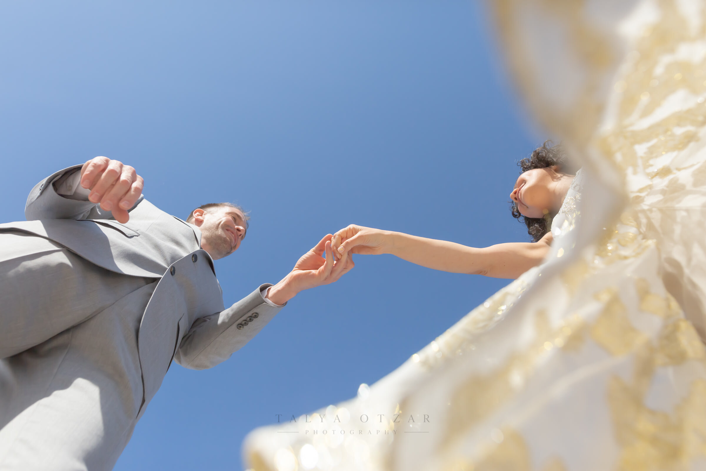 The groom reaches down to take the bride's hand, photographed from below against an open sky