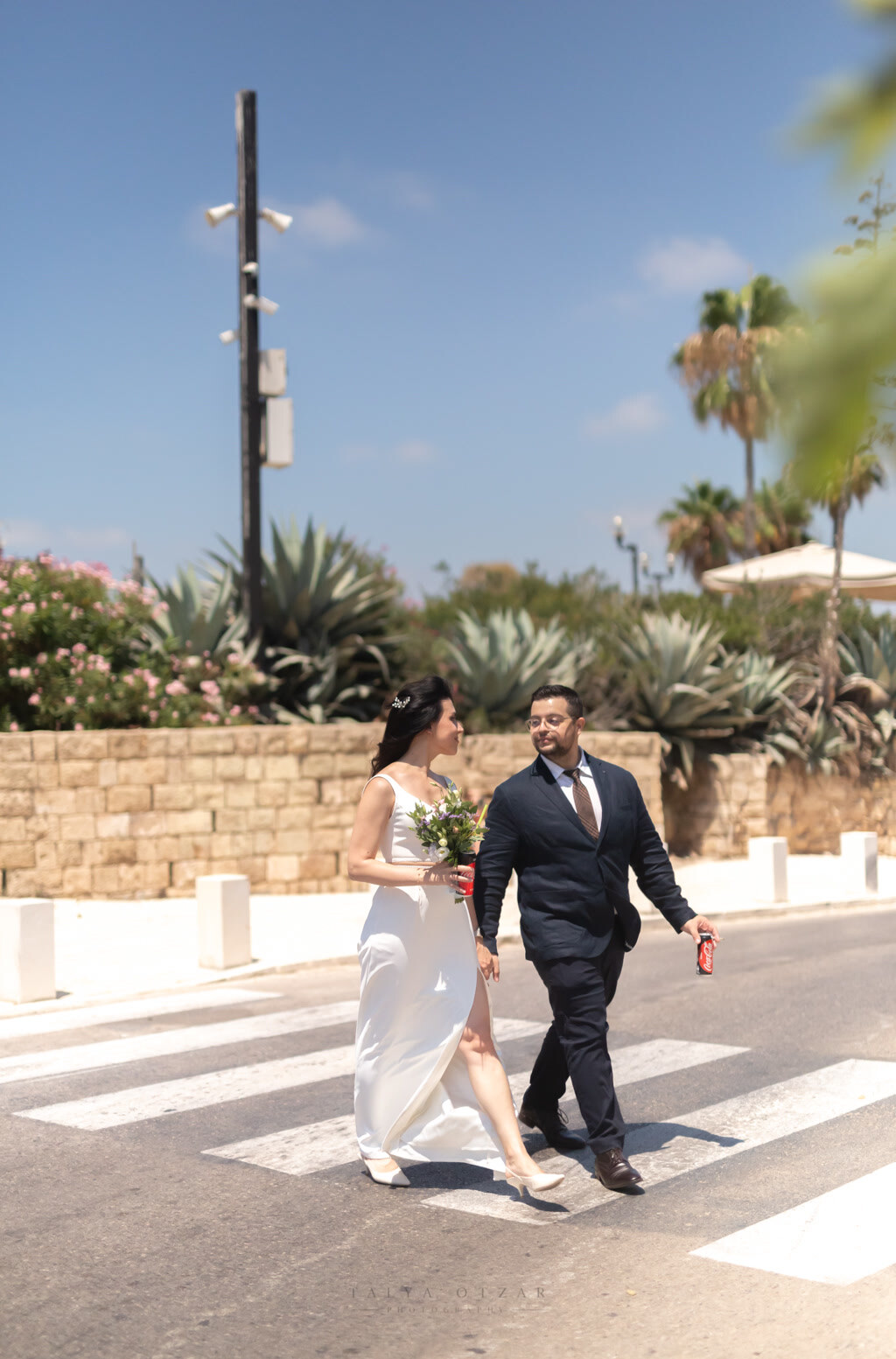 Bride and groom crossing a sunlit street, she carries a small bouquet and he a cold drink, agaves and palms behind