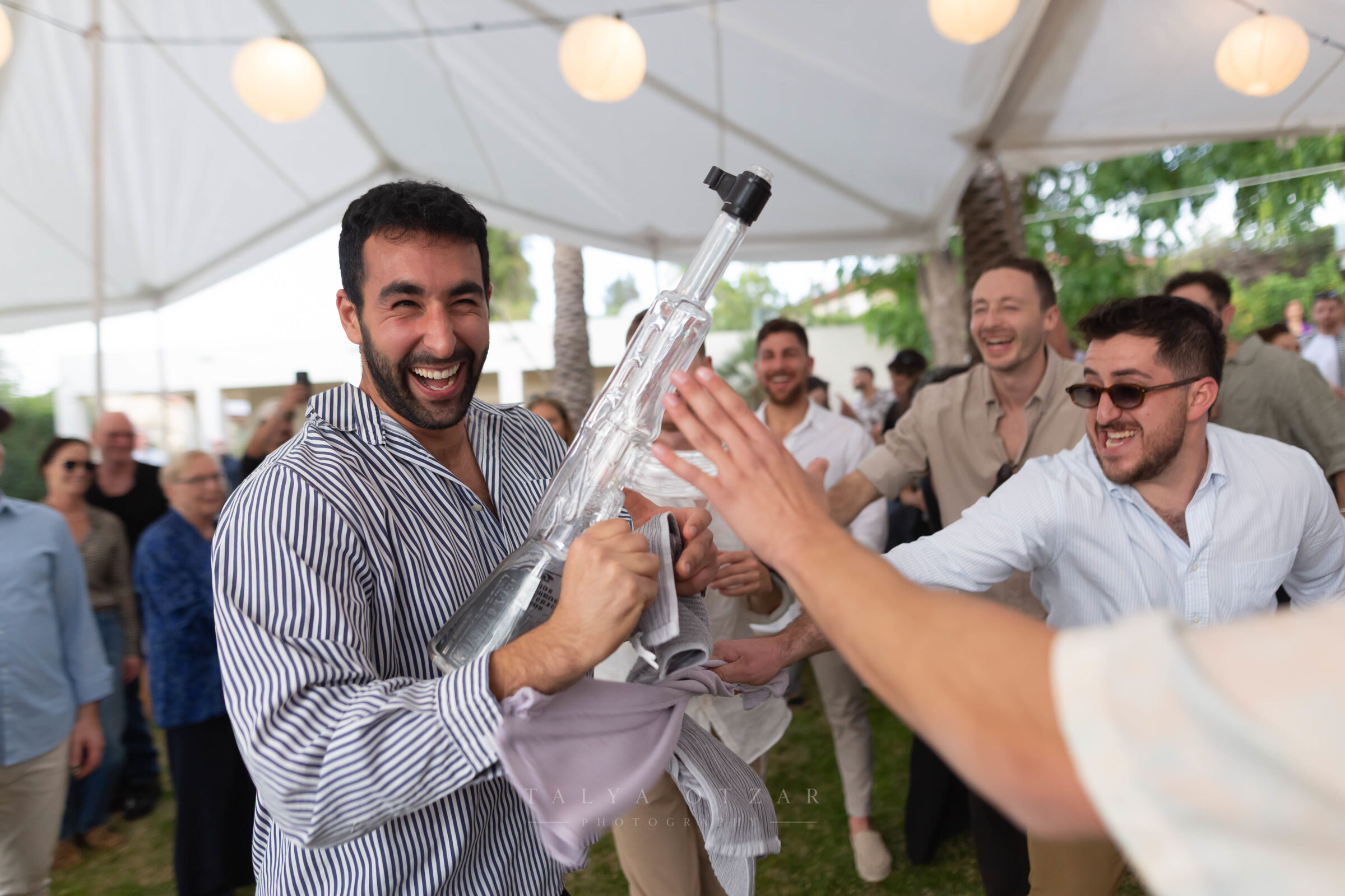 A guest laughing as friends crowd round him under the marquee, paper lanterns strung overhead