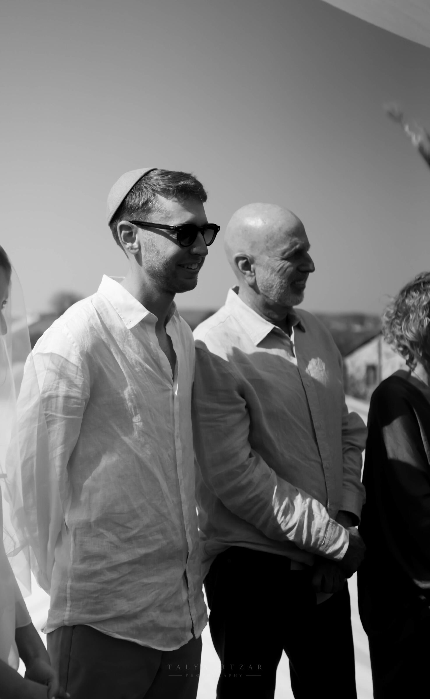 The groom in a kippah standing beside his father in the sun, the bride's veil catching the edge of the frame