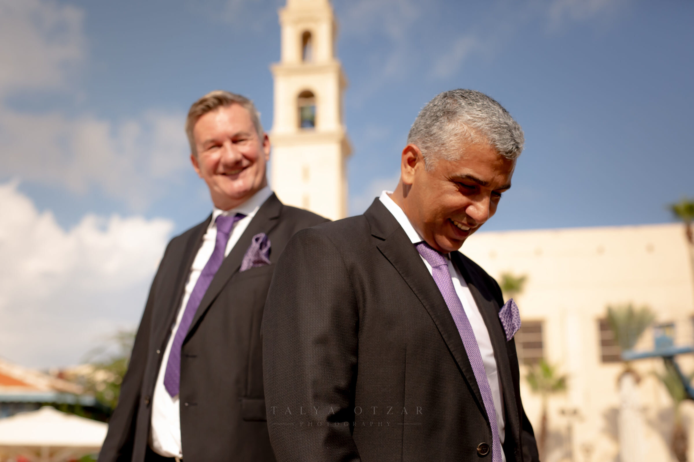 Two guests in dark suits and lilac ties outside in Jaffa, the church bell tower rising behind them