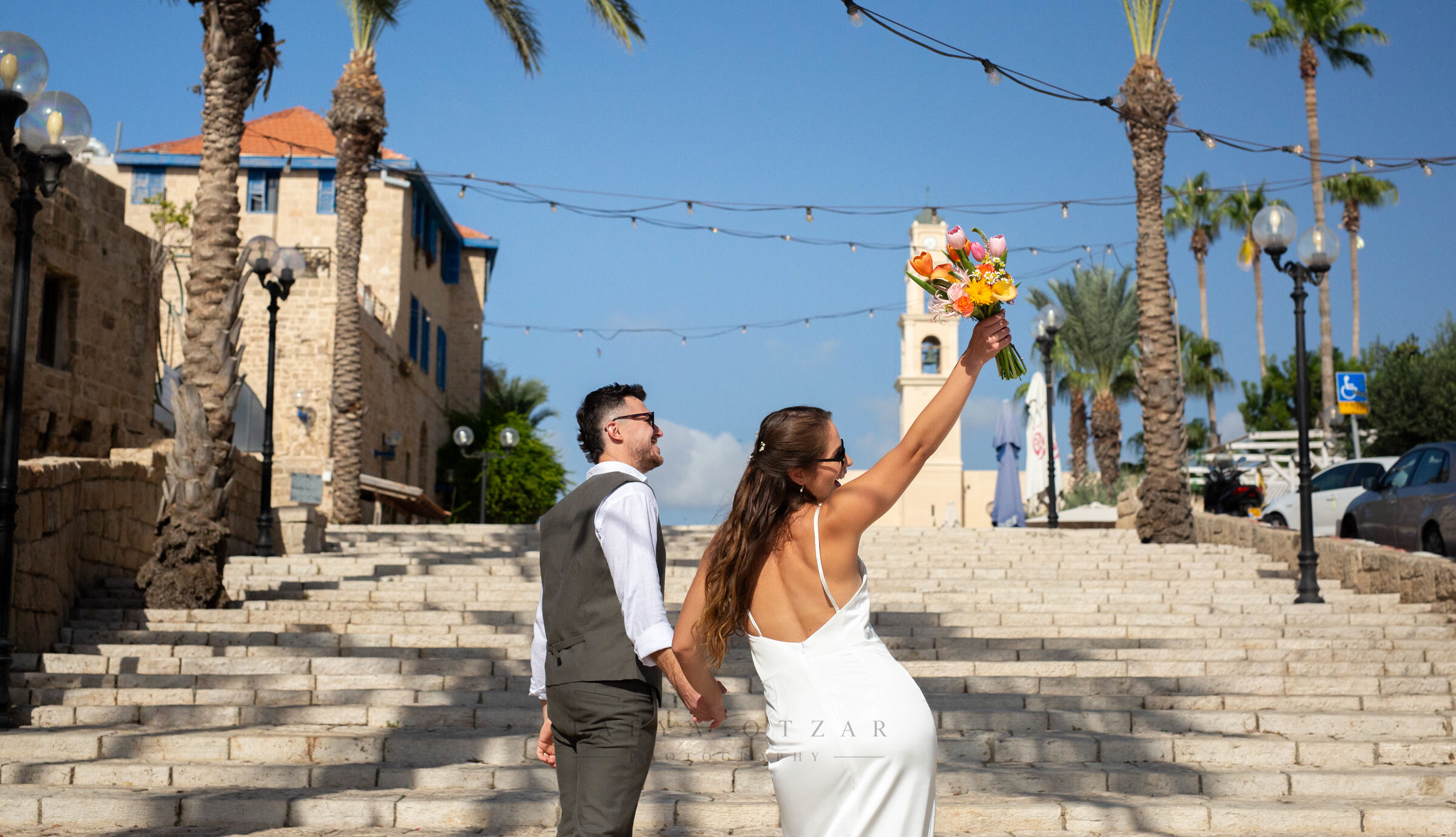The couple climbing the wide stone steps in Old Jaffa, the bride raising her bouquet above her head