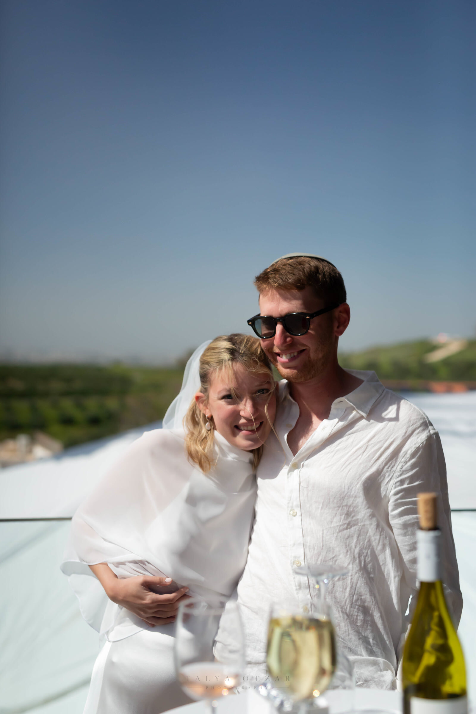 Bride and groom leaning into each other at an outdoor table, open countryside stretching out behind them