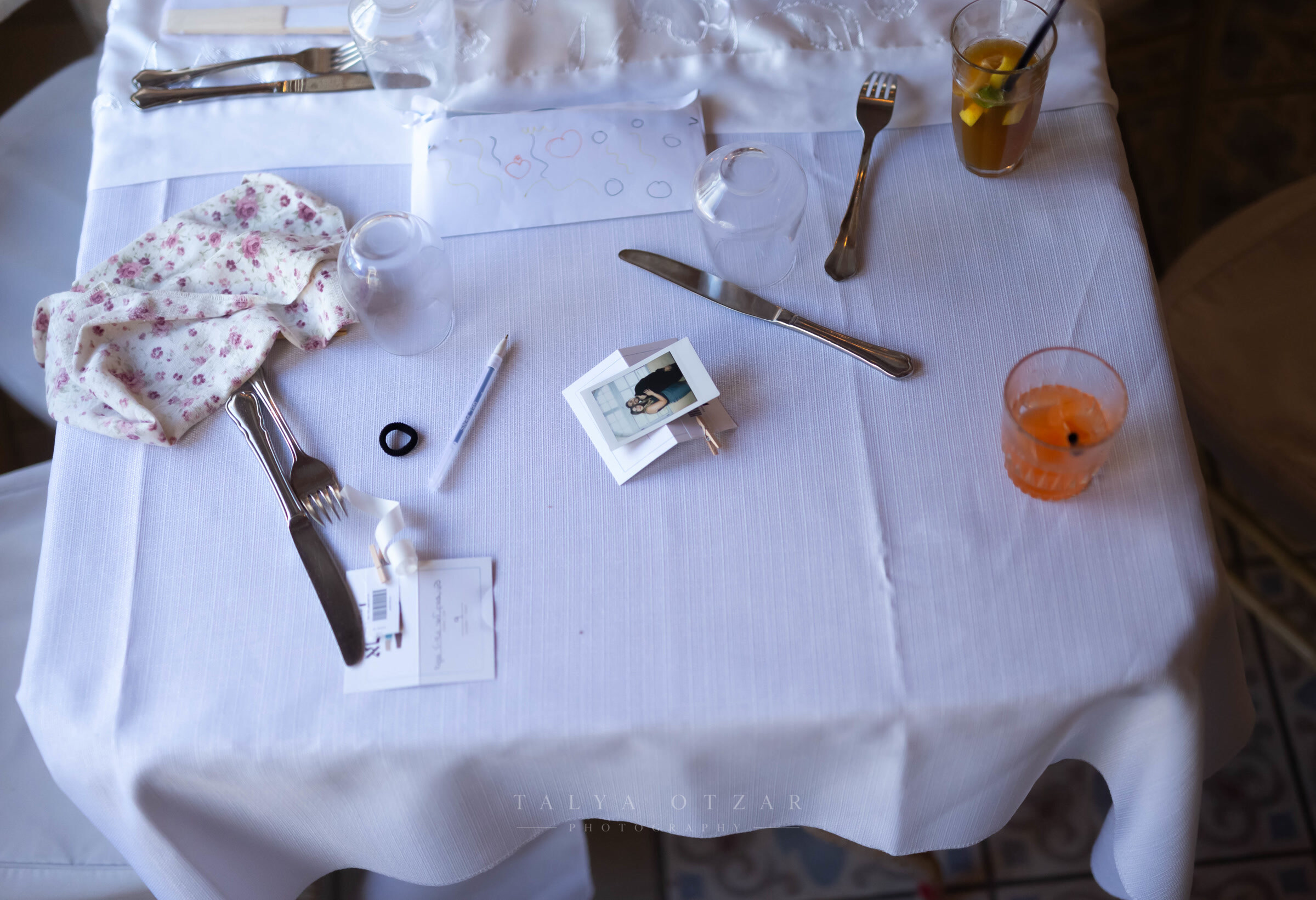 A corner of the wedding table from above — floral napkin, cutlery, an instant photo and two cold drinks