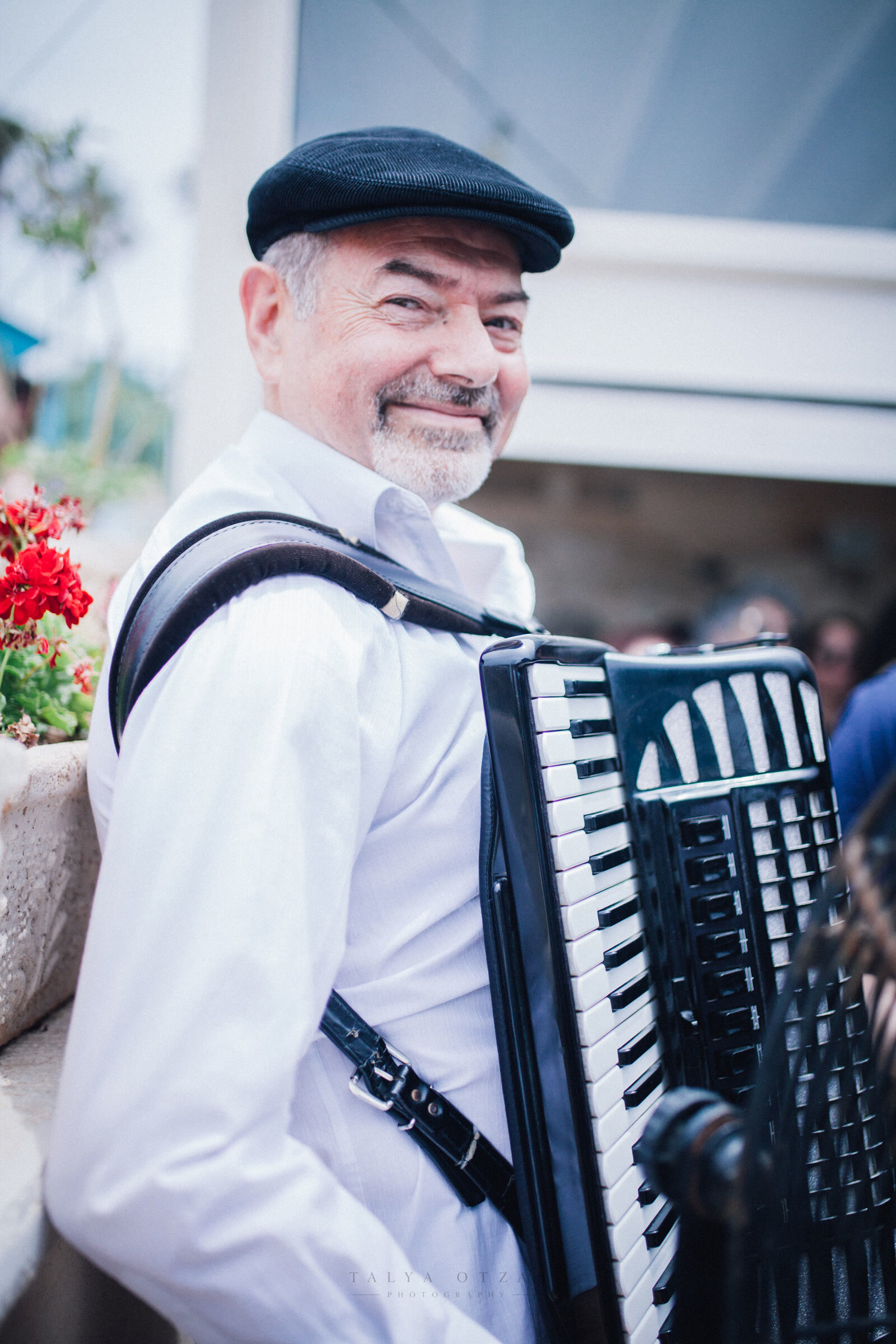 The accordion player smiling mid-tune, red geraniums flowering beside him