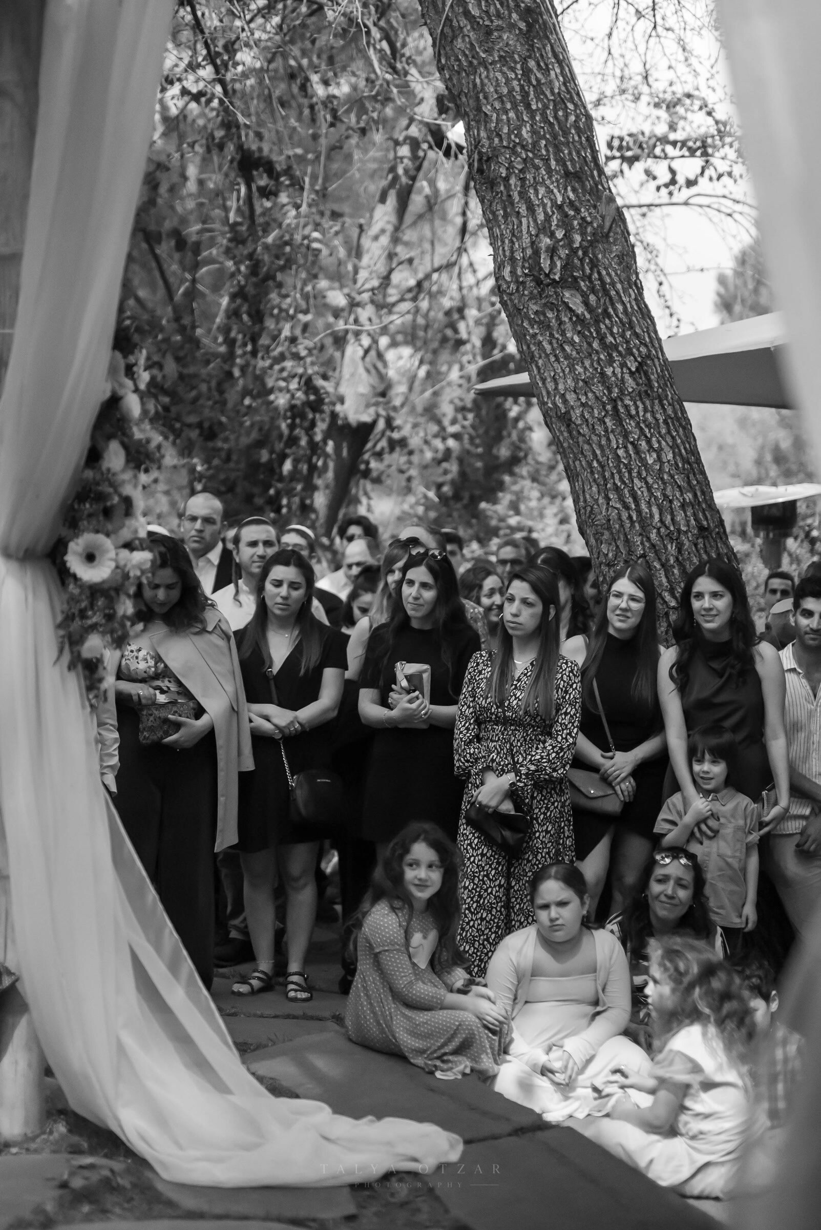 Guests standing under the trees to watch the ceremony, the bride's train and the chuppah drape in the foreground