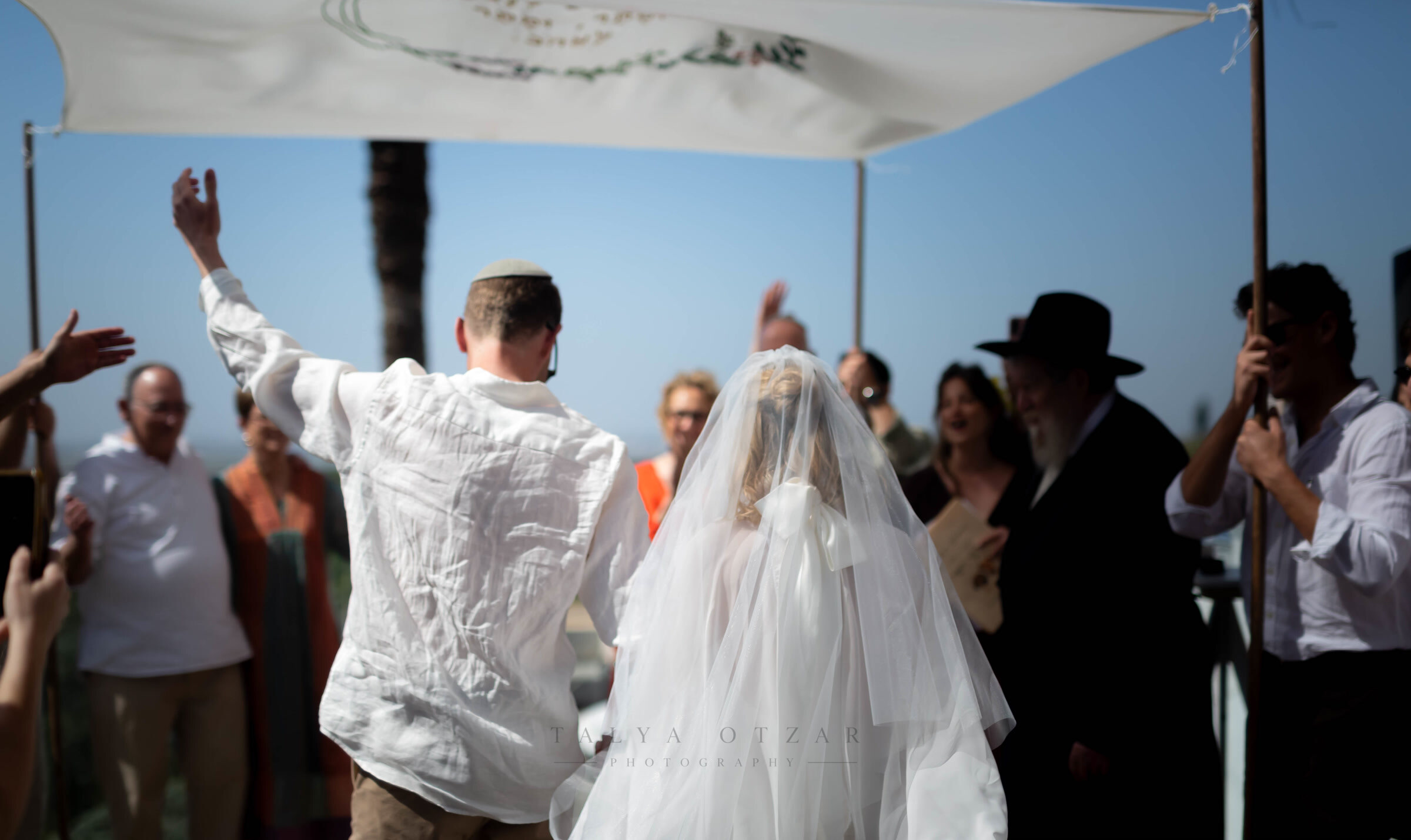 The couple walking out from under the tallit as guests clap, the sea behind them