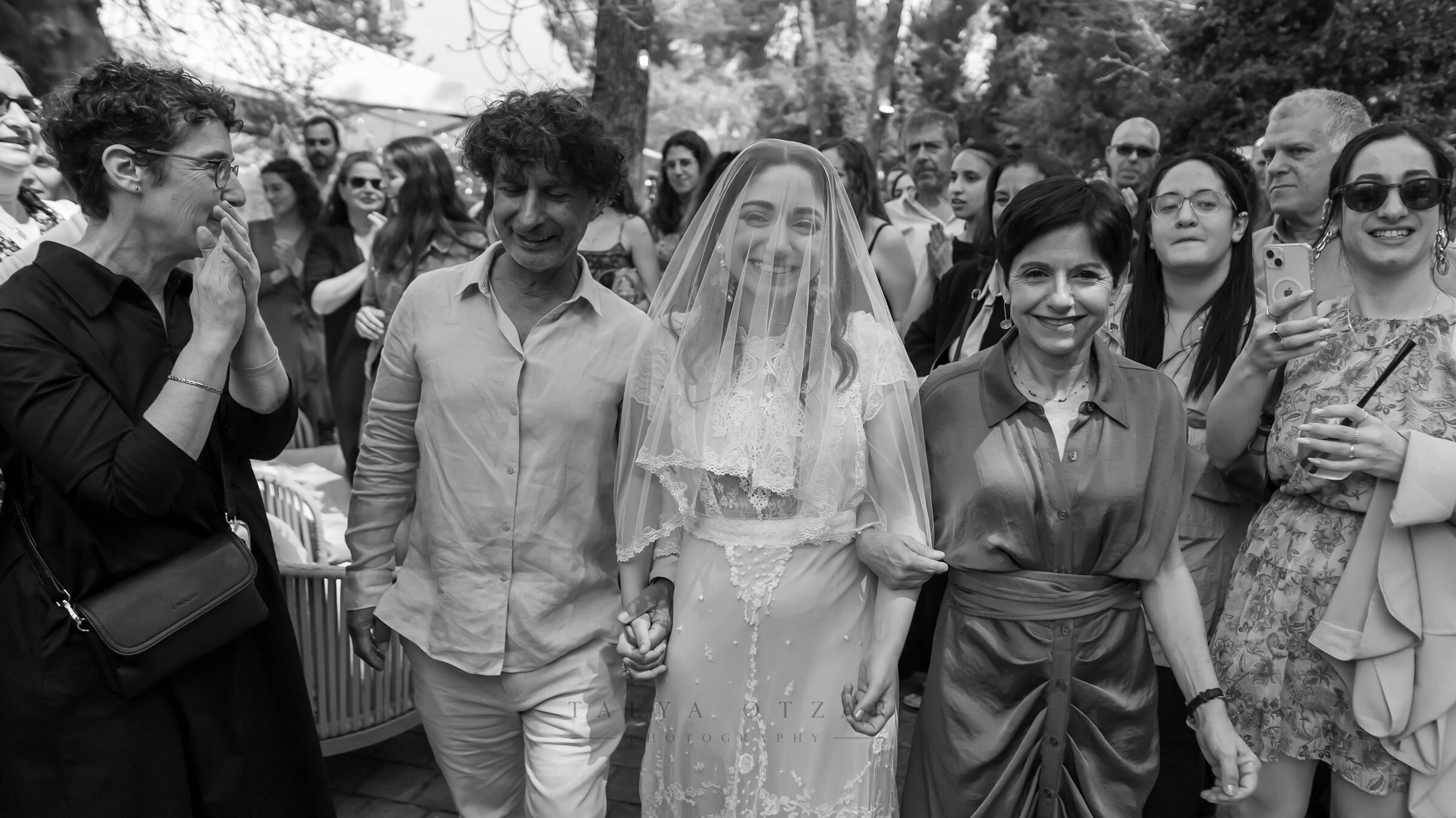 The veiled bride walking towards the chuppah between her partner and her mother, guests smiling on either side