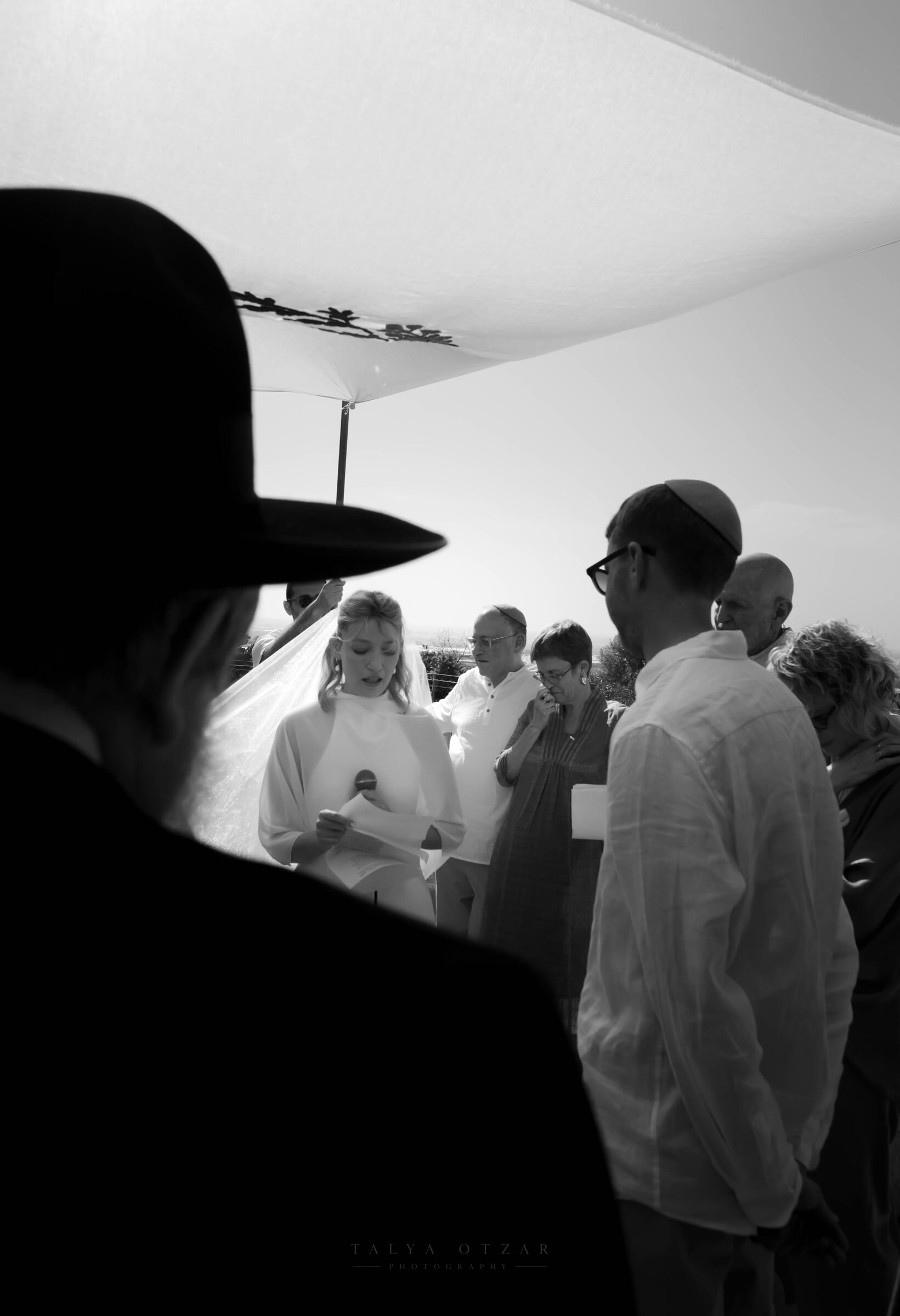 The bride reading her vows aloud under the chuppah, the rabbi's hat dark in the foreground