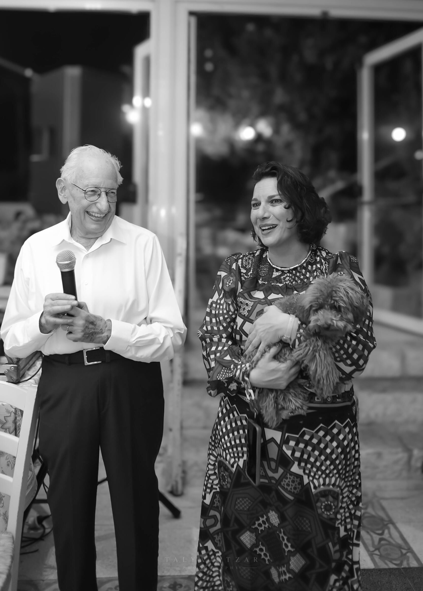 The grandfather laughing mid-speech, microphone in hand, a small dog held beside him