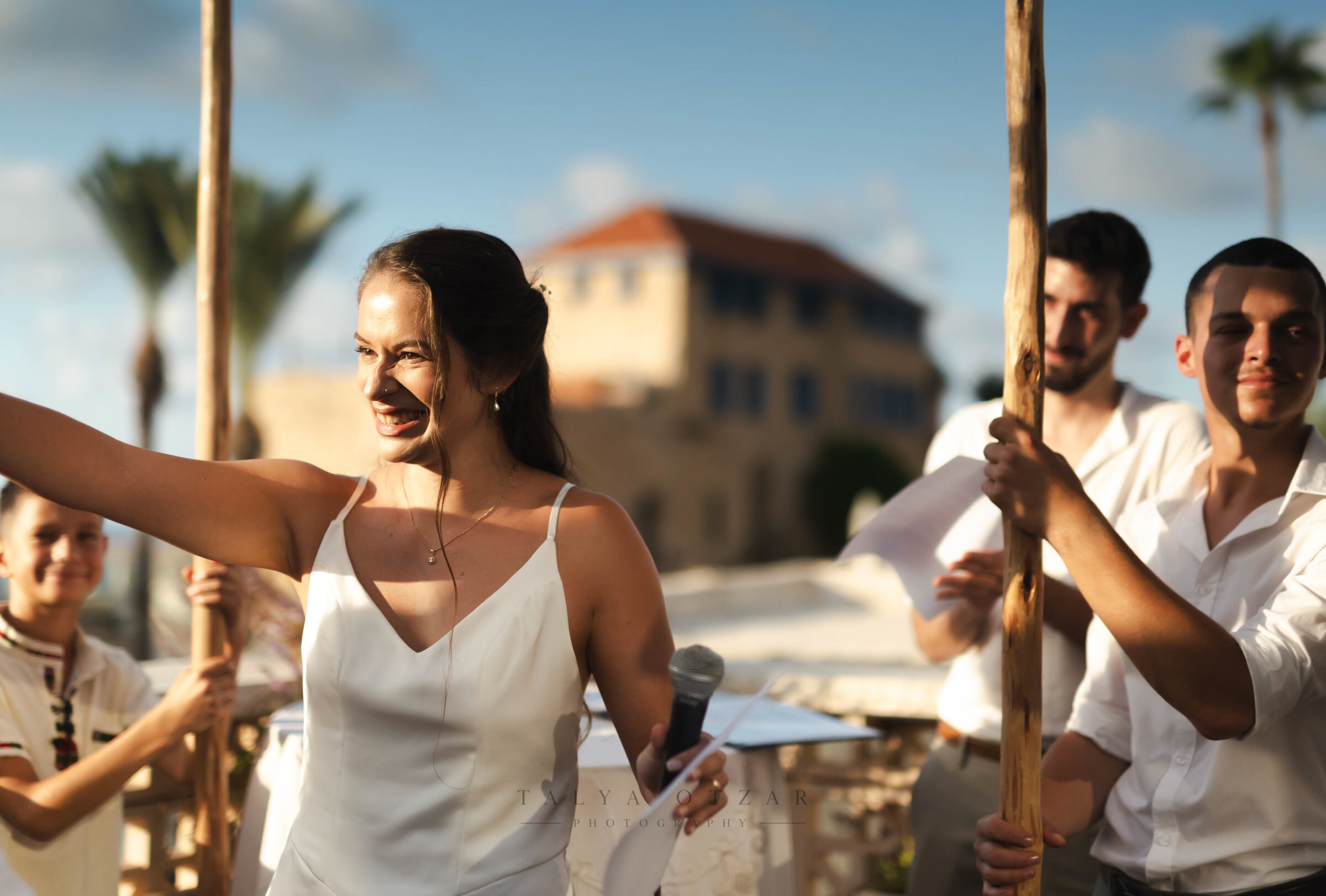 A guest laughing into a microphone while the chuppah poles are carried in behind her