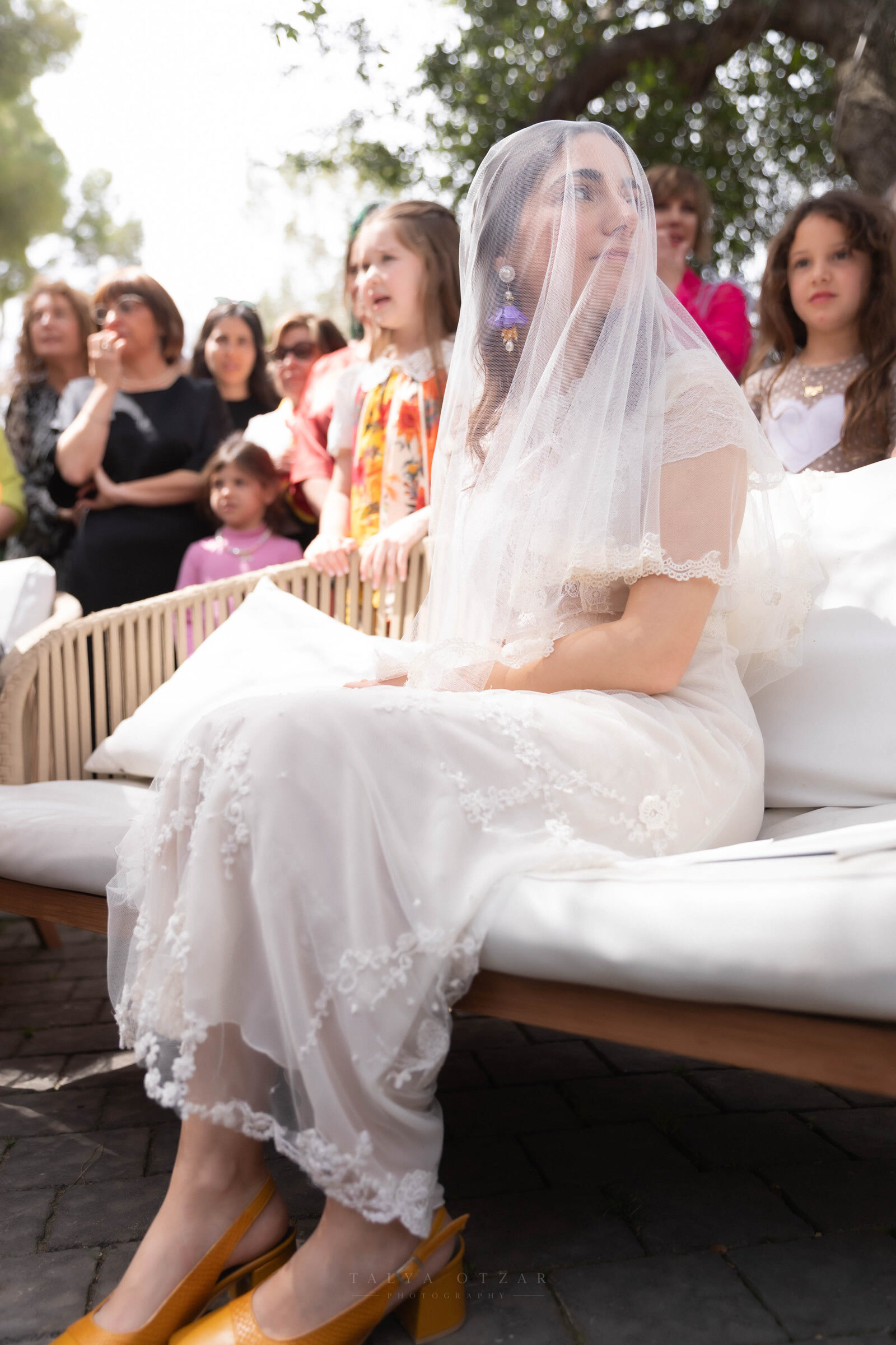 The bride waiting alone on a bench before the ceremony, her veil over her face and guests behind