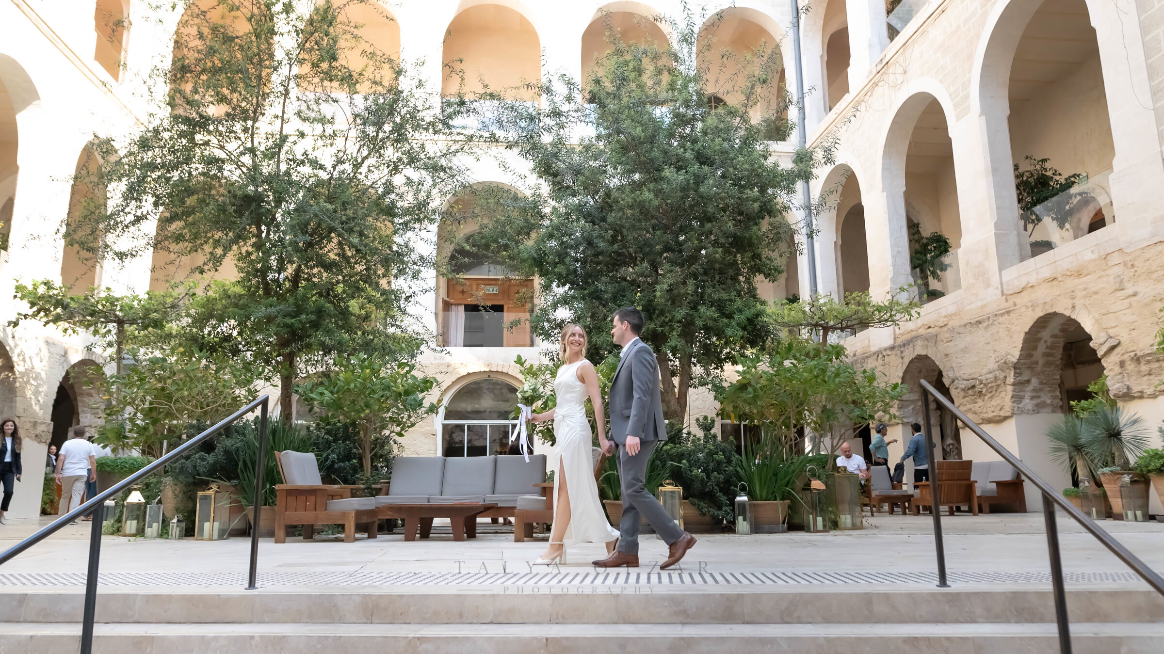 Bride and groom crossing an arcaded stone courtyard planted with olive trees