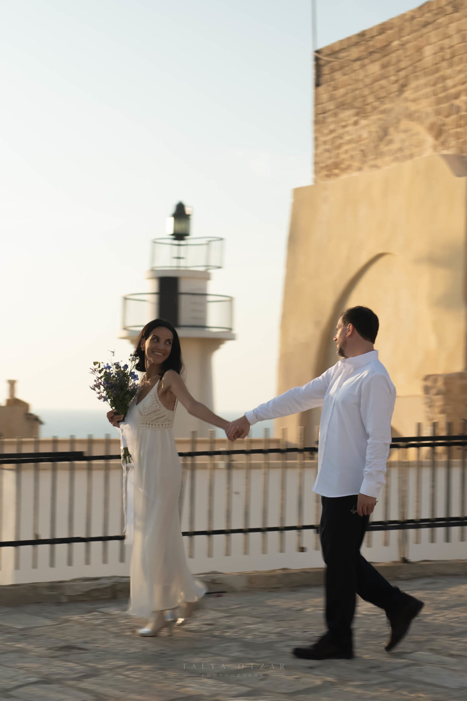 The couple walking the seafront path at sunset, an old lighthouse and stone fort behind them