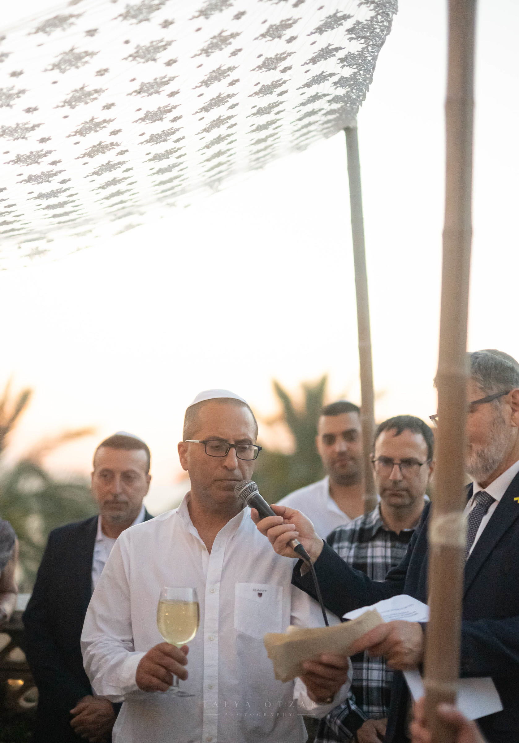 A blessing read aloud under the lace chuppah at sunset, a glass of wine in hand