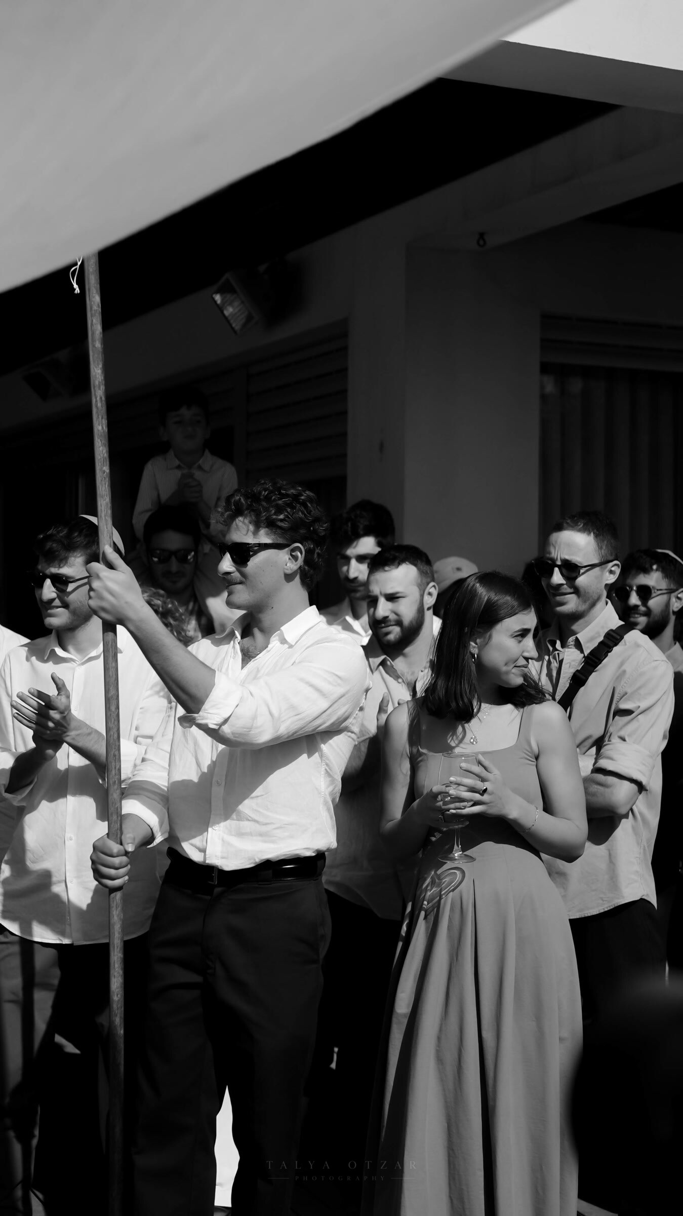 A friend holding one of the chuppah poles as the guests applaud in the sun