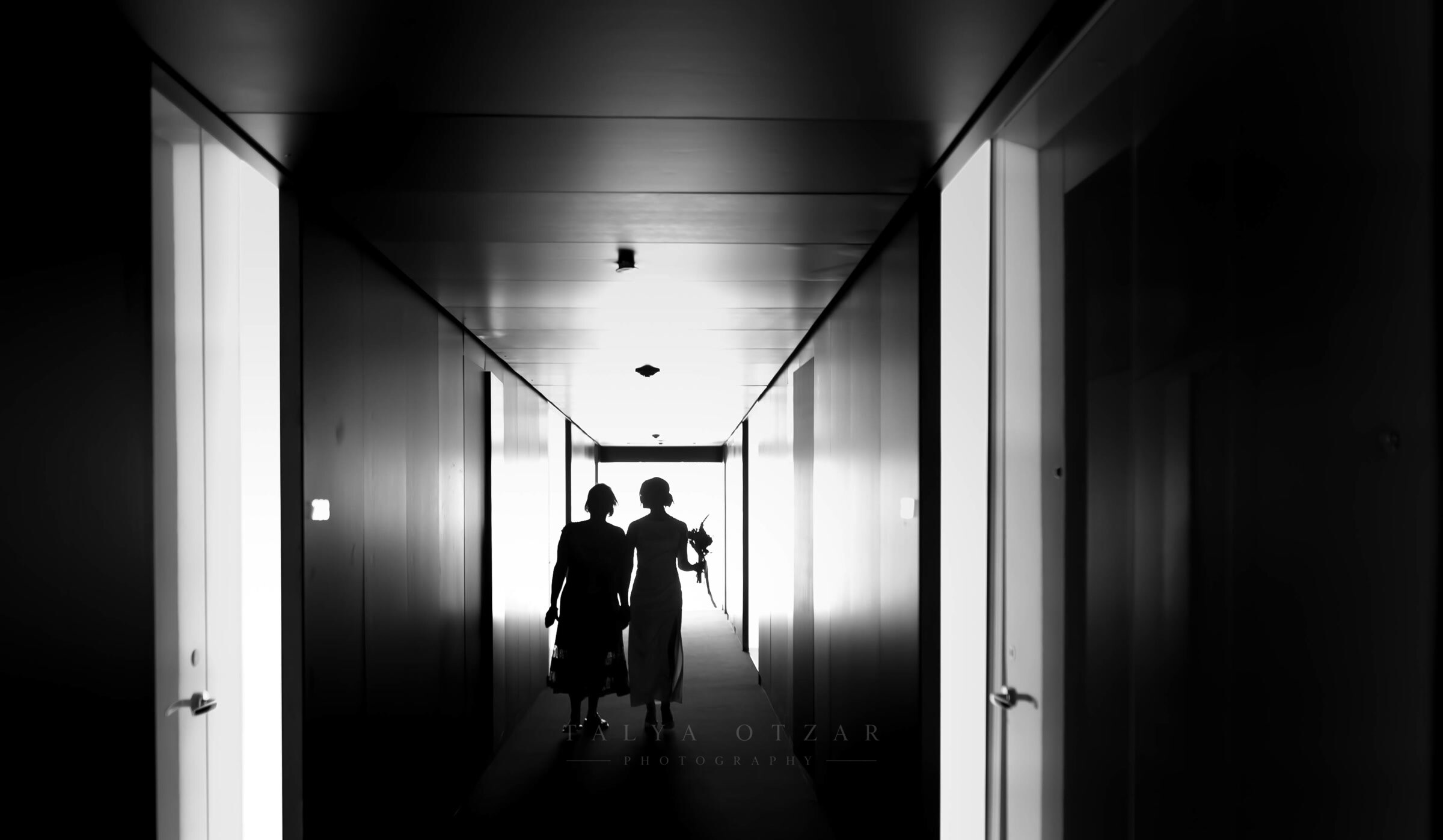 The bride and her mother silhouetted at the end of a dark hotel corridor, walking towards the light