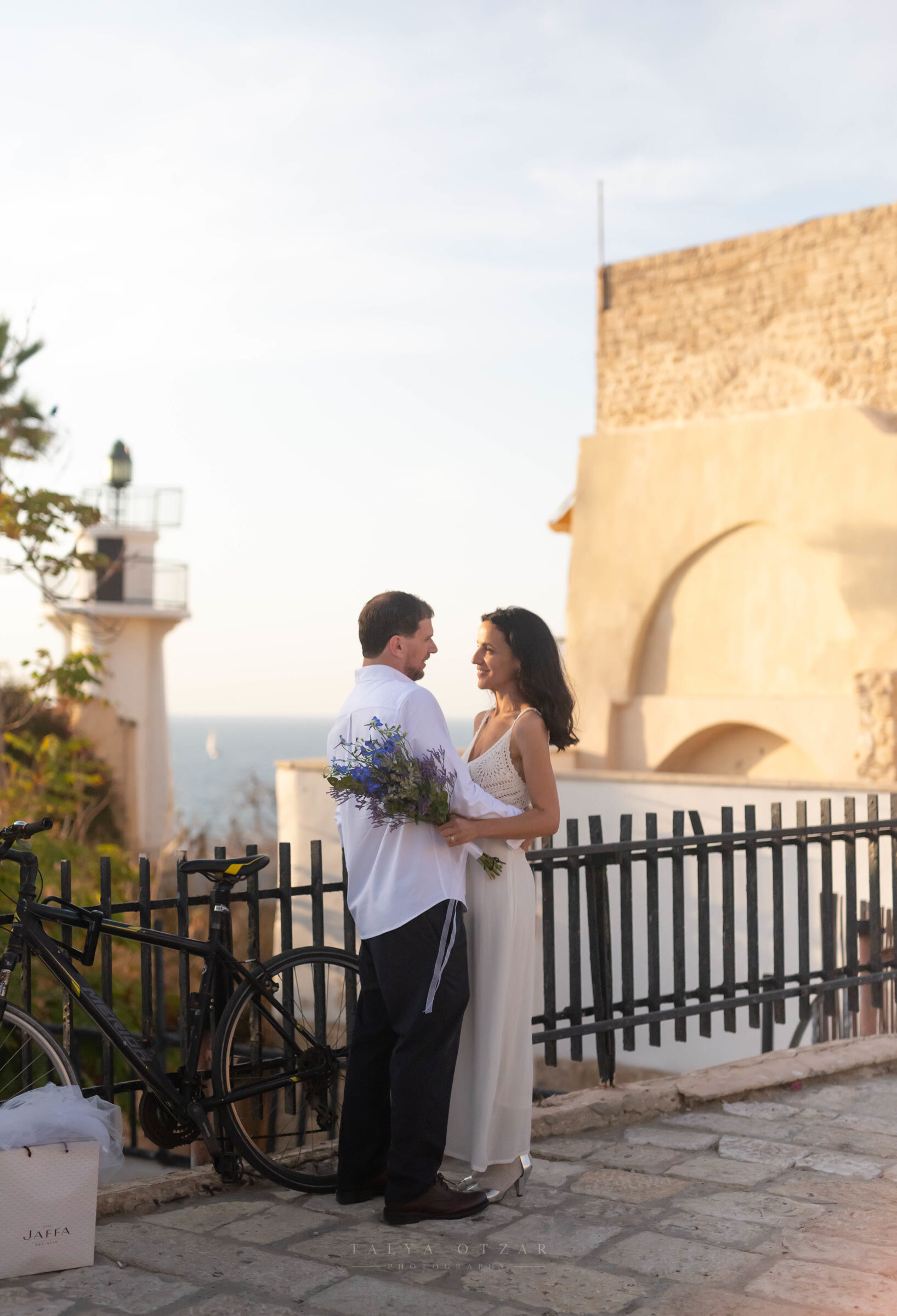 The couple laughing face to face on the Jaffa clifftop at golden hour, the lighthouse and sea behind