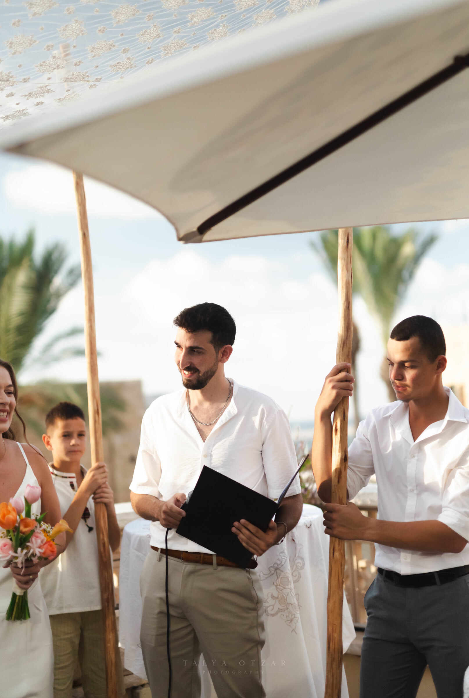 Two friends holding the chuppah poles as the officiant reads, the sea and palms behind them