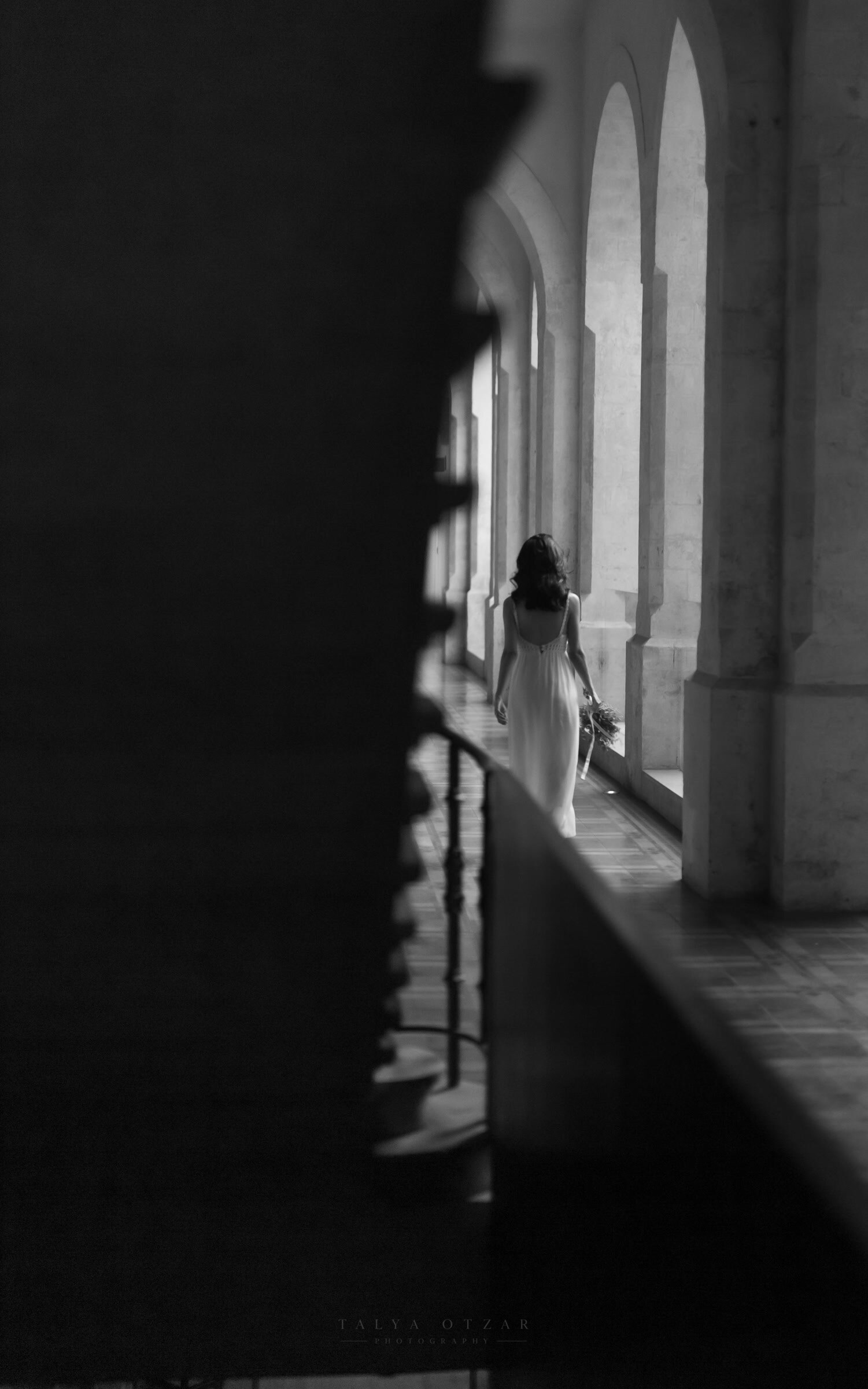 The bride walking away down an arched stone cloister, bouquet in hand