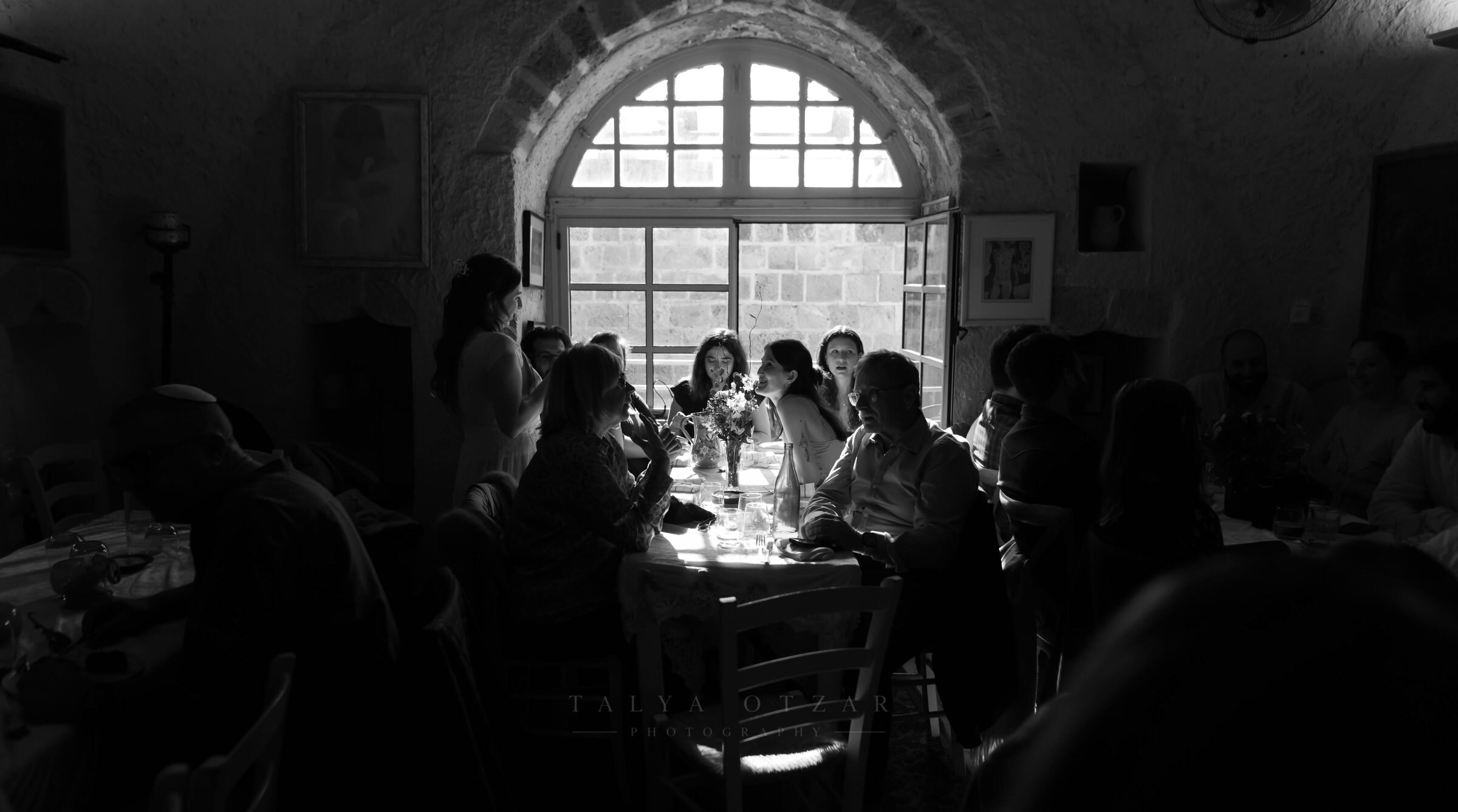 Guests around the long table in the vaulted stone room, lit only by the arched window