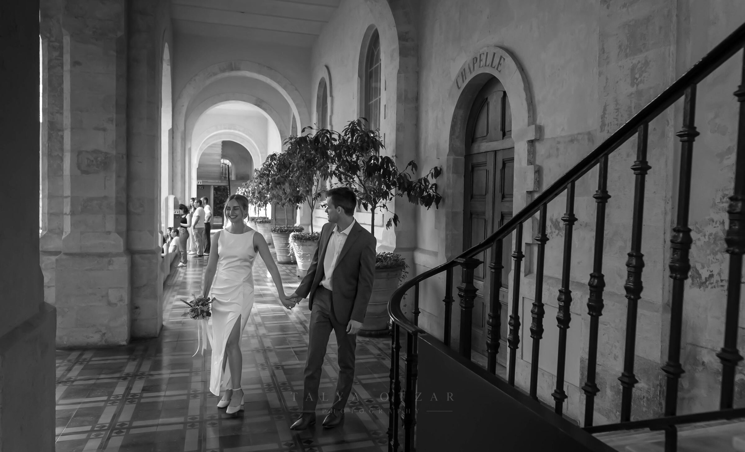 The couple walking a colonnaded corridor past a chapel doorway
