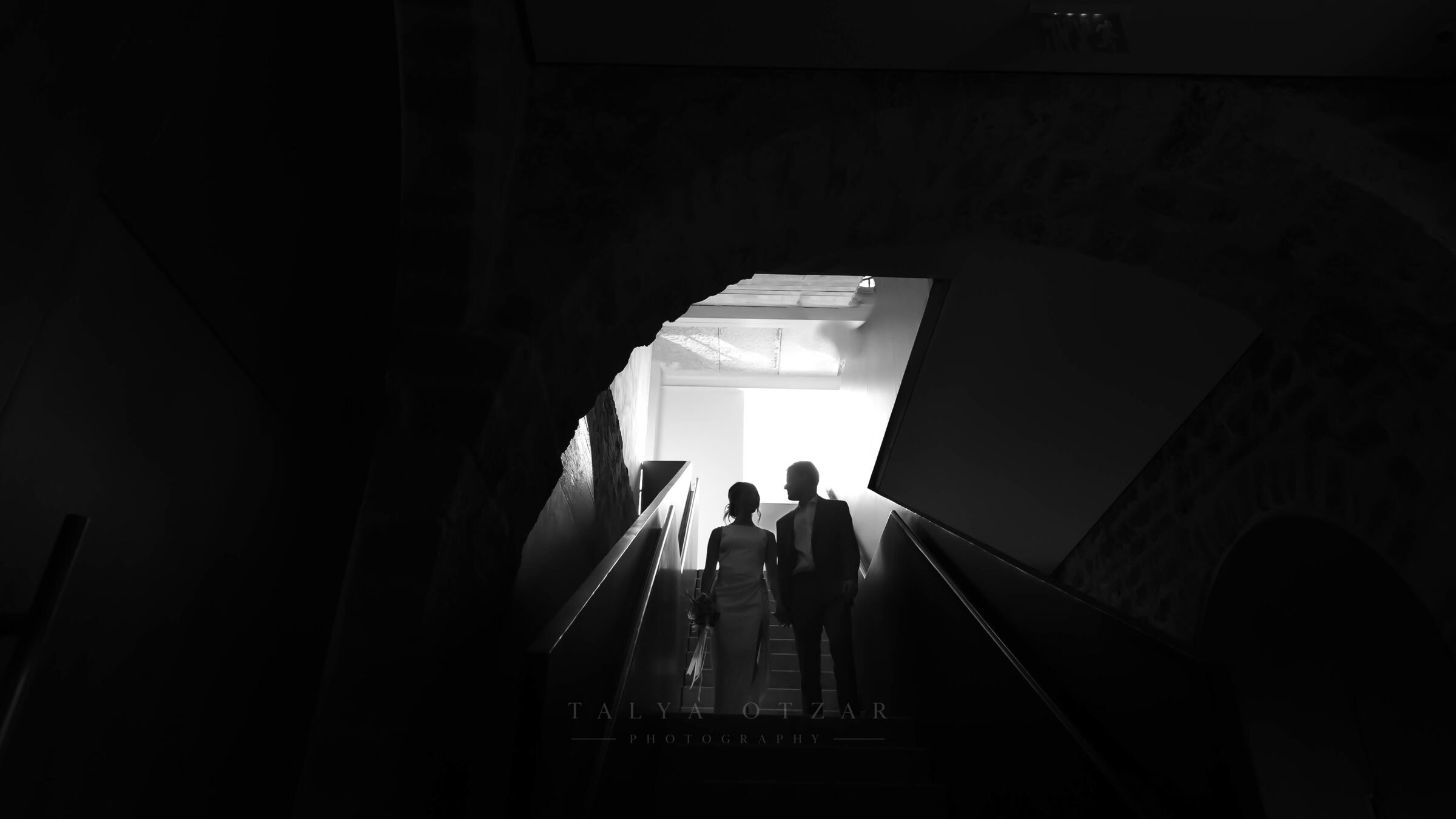 Bride and groom silhouetted at the top of a dark staircase, stepping into the light