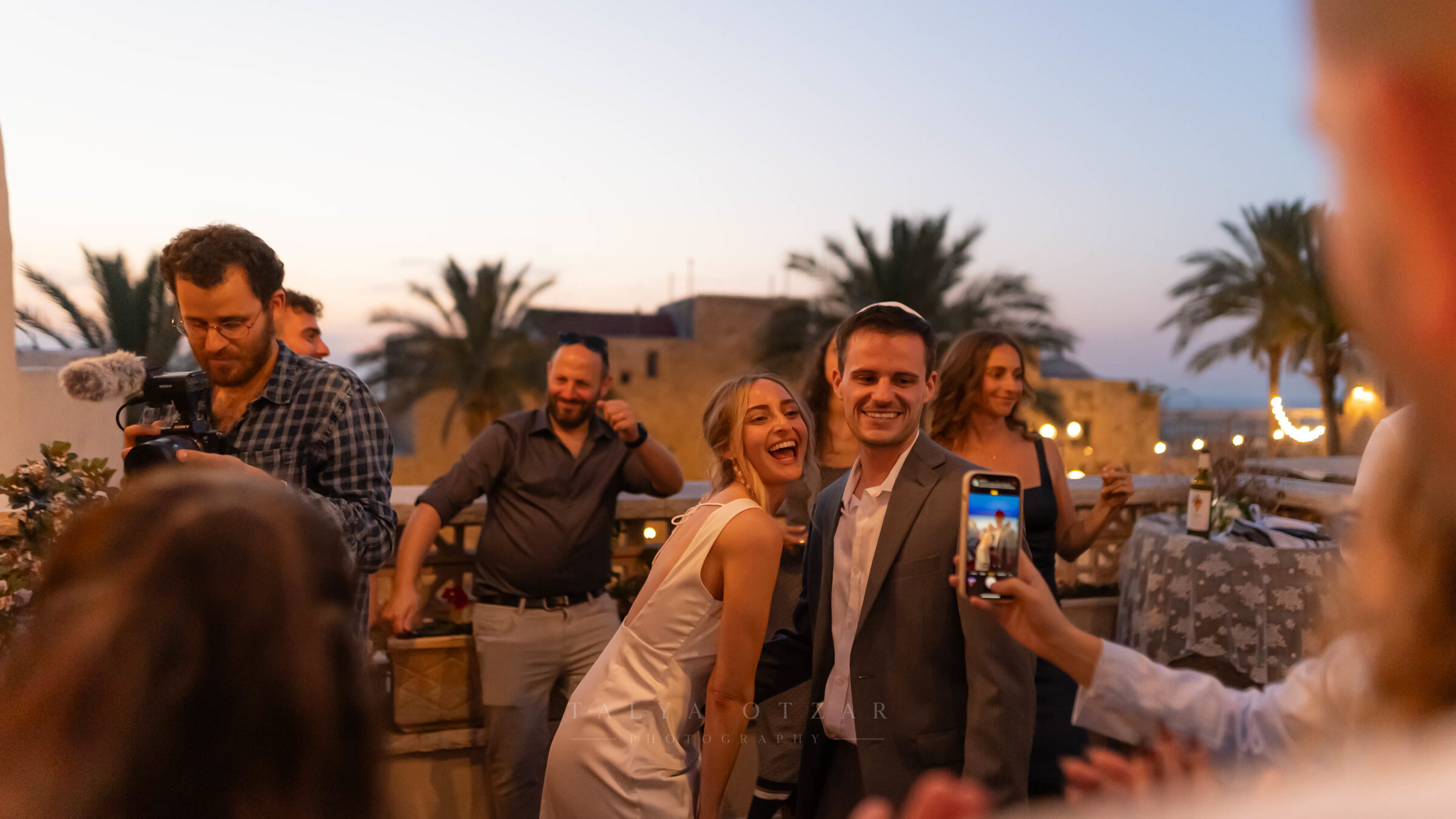 The couple laughing on the Jaffa rooftop at dusk while a guest films them on a phone