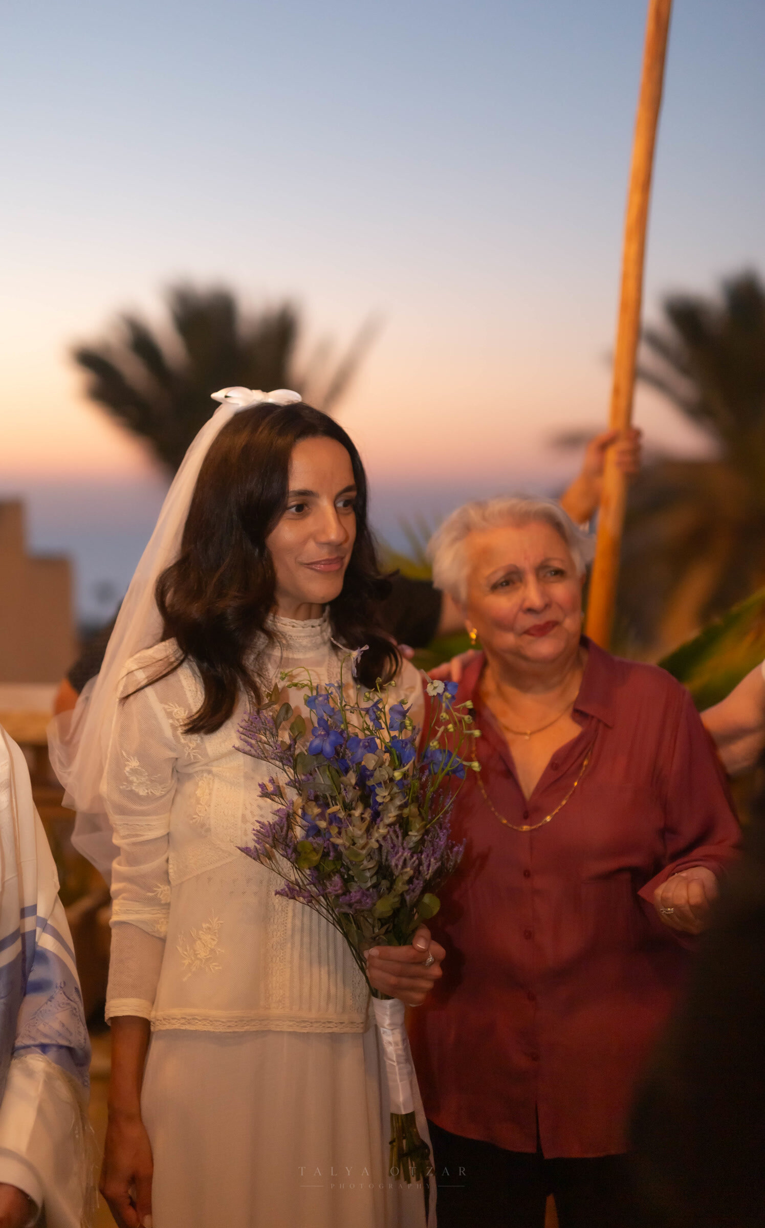 The bride in a high-necked lace dress carrying blue wildflowers, walking beside her grandmother at sunset