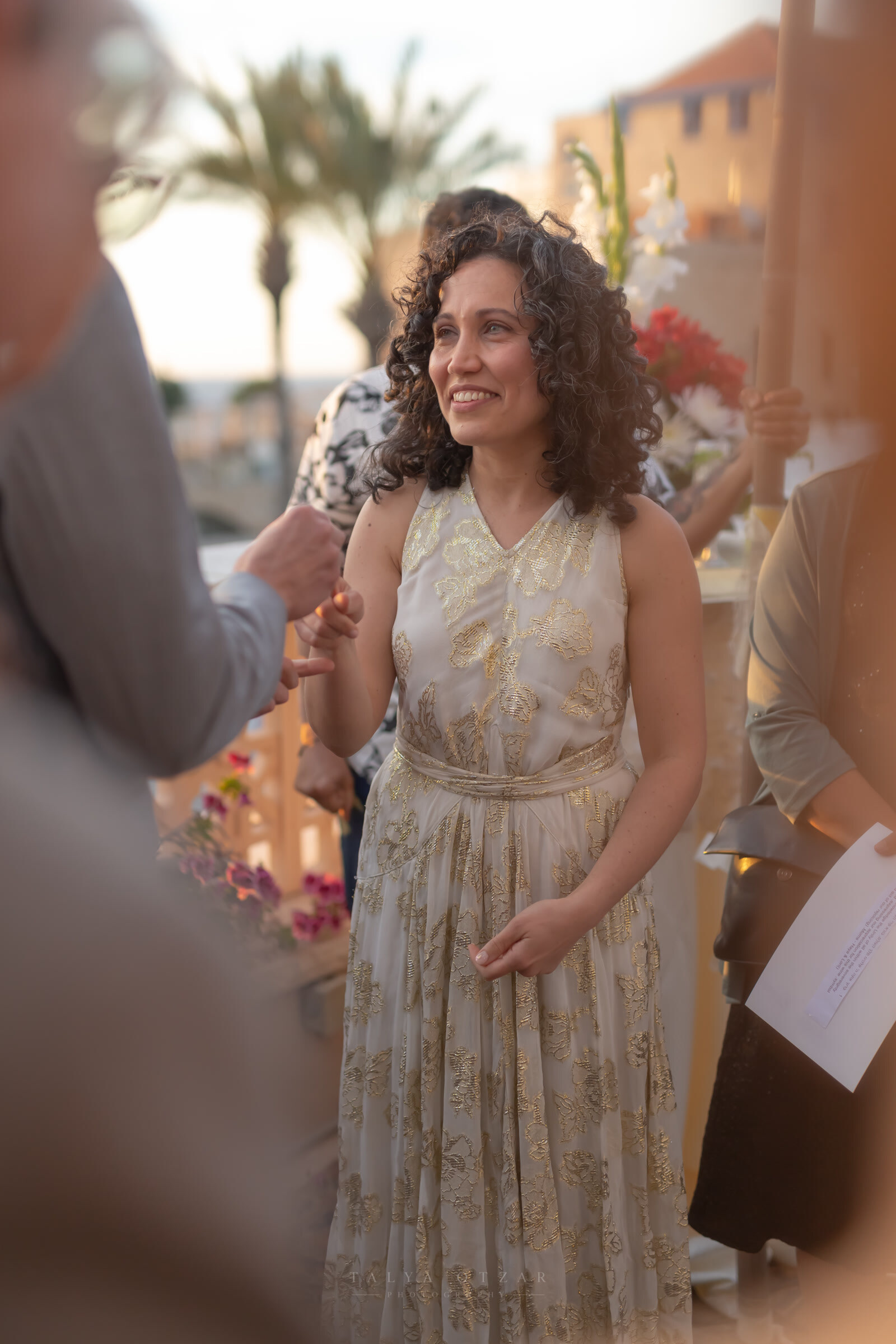 A guest in a gold-embroidered dress smiling through the ceremony, candles and flowers behind her